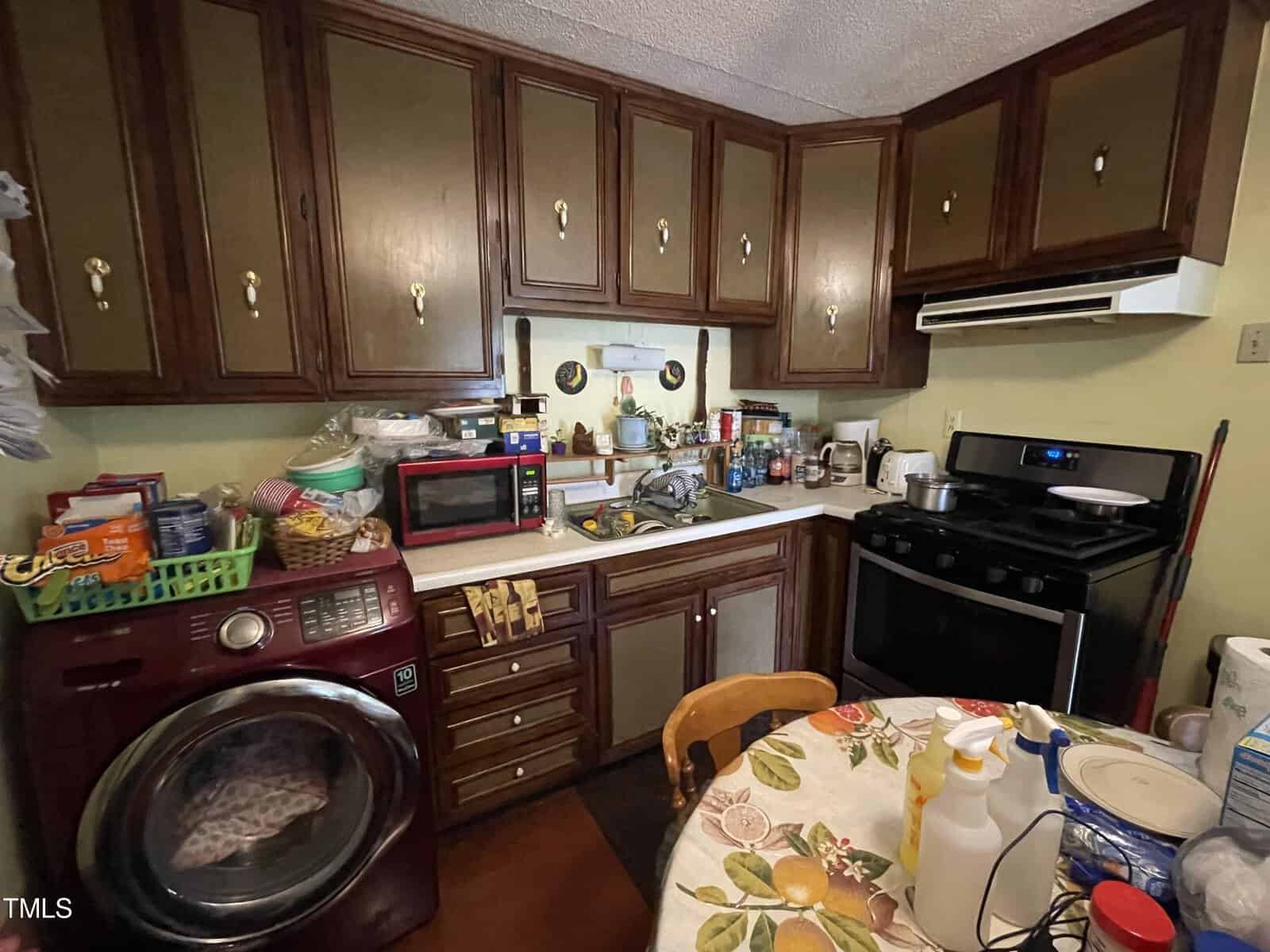 A cluttered kitchen with dark wooden cabinets, a variety of small appliances on the counters, and a dining table with a floral tablecloth. there's also a washing machine on the left side.