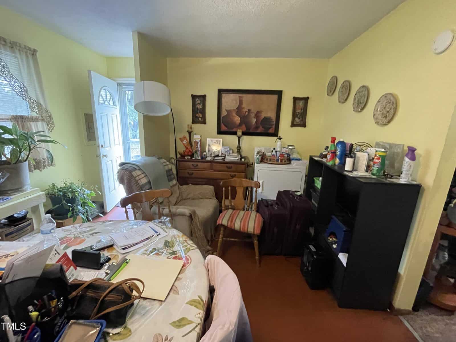 A cluttered living room with a yellow wall, decorated with various wall hangings, plants, and a mix of furniture including a desk covered with paperwork and items.