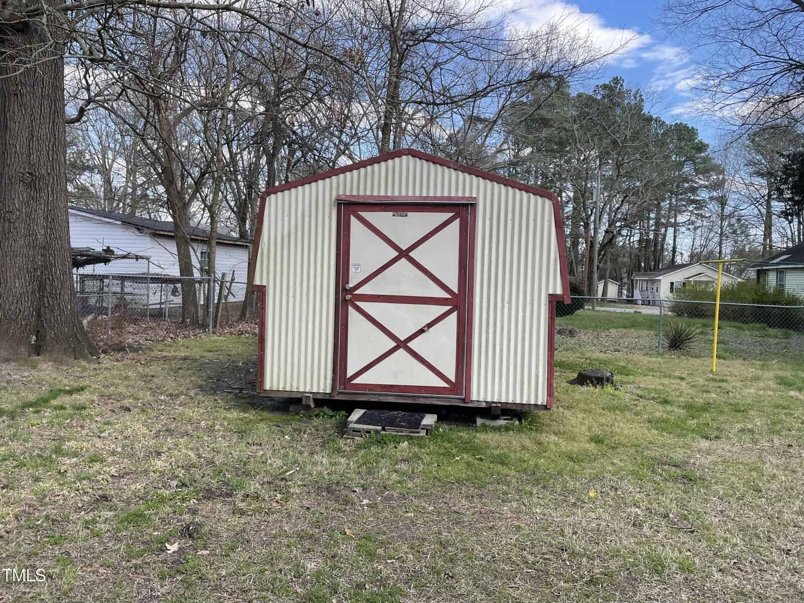 A small, white and red mobile home with a barn-style door, positioned in a grassy yard surrounded by trees and a residential neighborhood in the background.