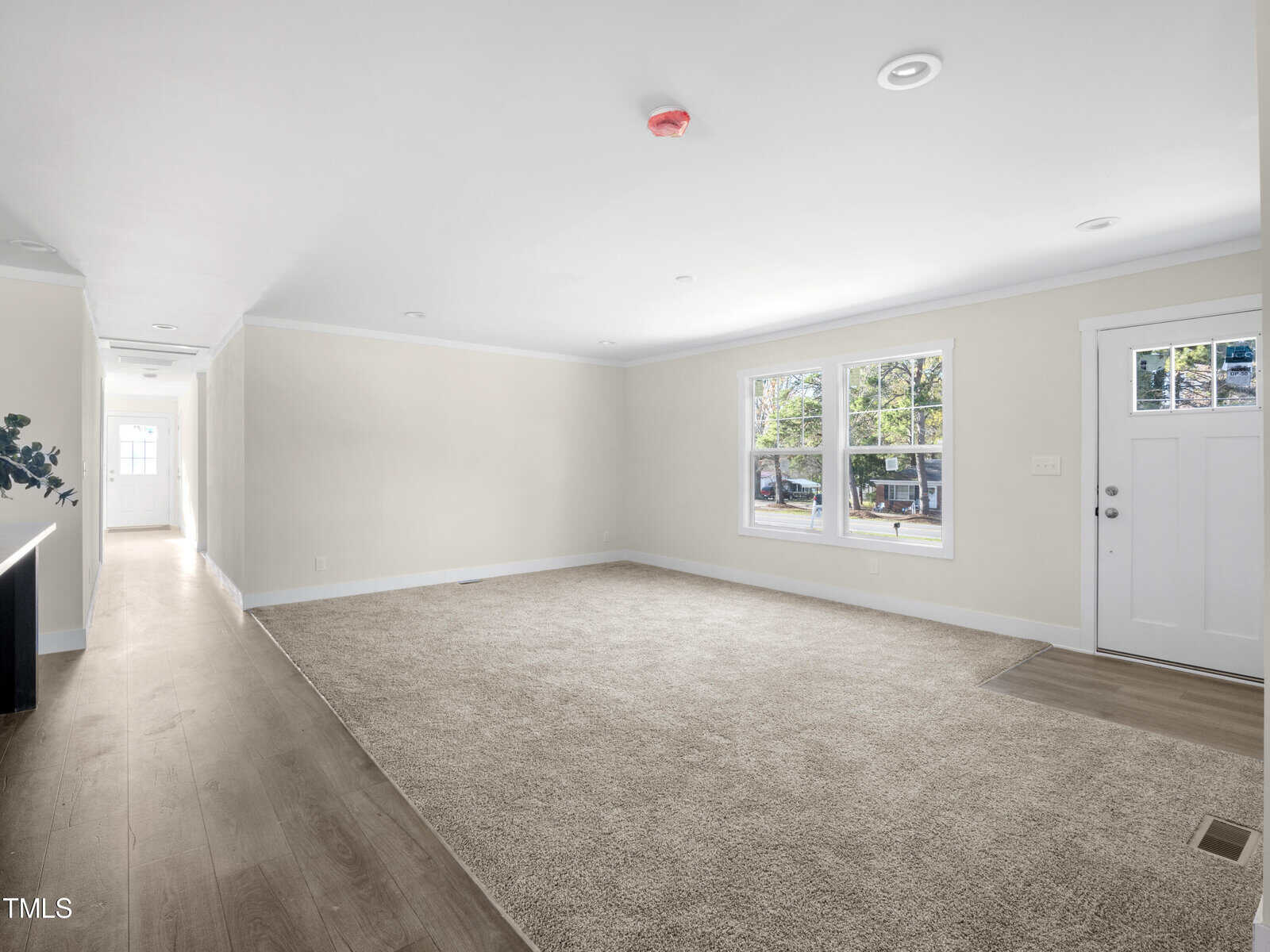 A spacious, unfurnished room with beige walls and carpeted flooring. There are two large windows letting in natural light and a white door. The room extends into a hallway with hardwood floors and a small table with a plant on the left side.