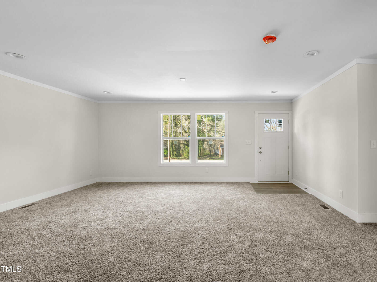 A spacious, unfurnished living room with beige carpet and white walls. Two large windows and a door with a window provide ample natural light. A smoke detector is visible on the ceiling. The room is empty, showcasing its size and potential for various furnishings.