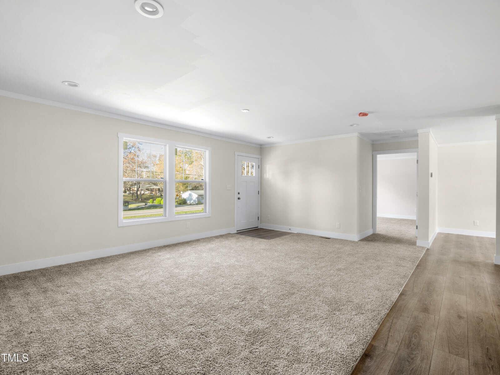 A spacious, empty living room with neutral-colored walls and beige carpeting. Two large windows let in natural light, and there is a white front door. The room transitions to a hardwood floor hallway that leads to an adjacent space.