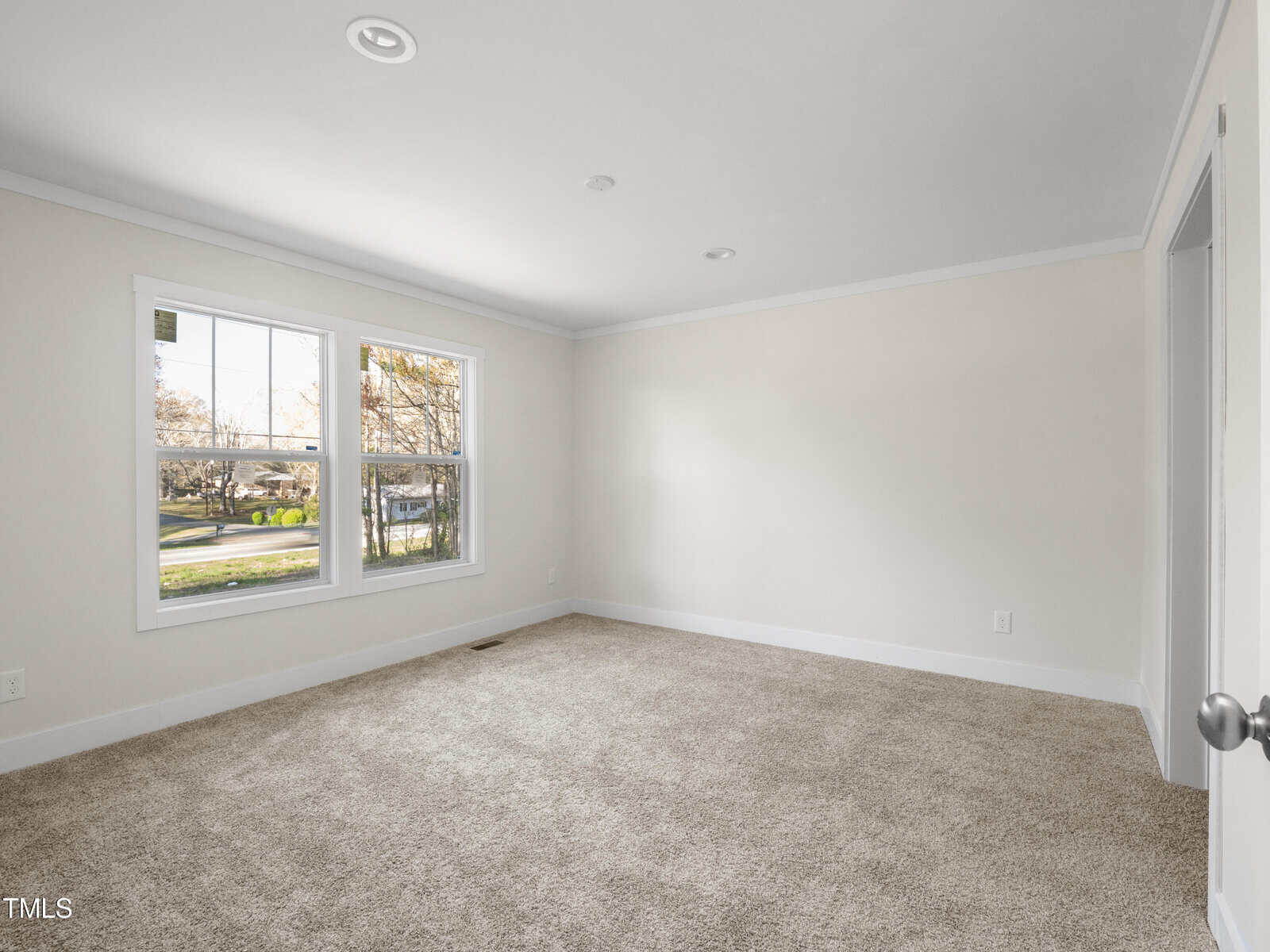 A bright, empty living room with beige carpet and light-colored walls. The room has large windows on the left side, allowing natural light to enter, and a door on the right side. The ceiling has recessed lighting, and the view outside shows a green yard.