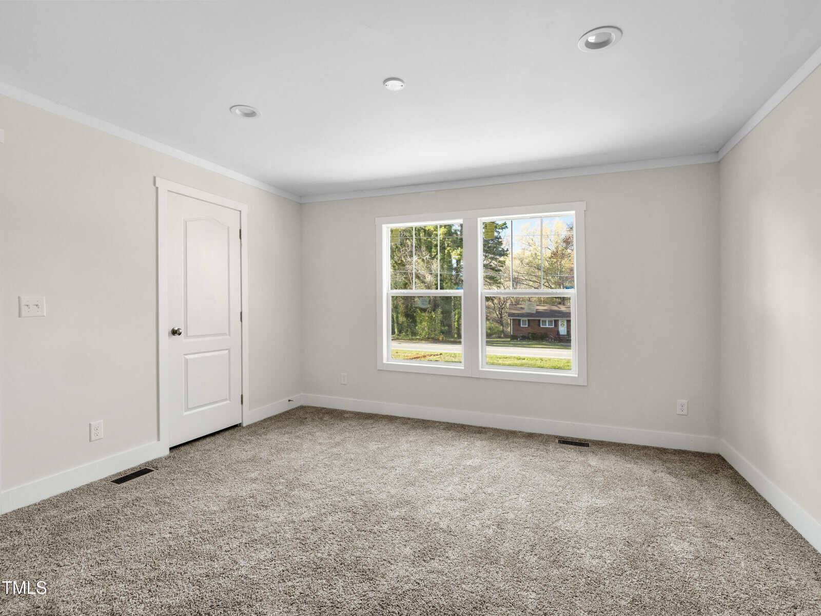 A bright, empty room with beige walls, a white ceiling, and light gray carpet. The room has a single white door on the left wall and a large double window on the far wall, allowing natural light to illuminate the space. A house can be seen outside through the window.