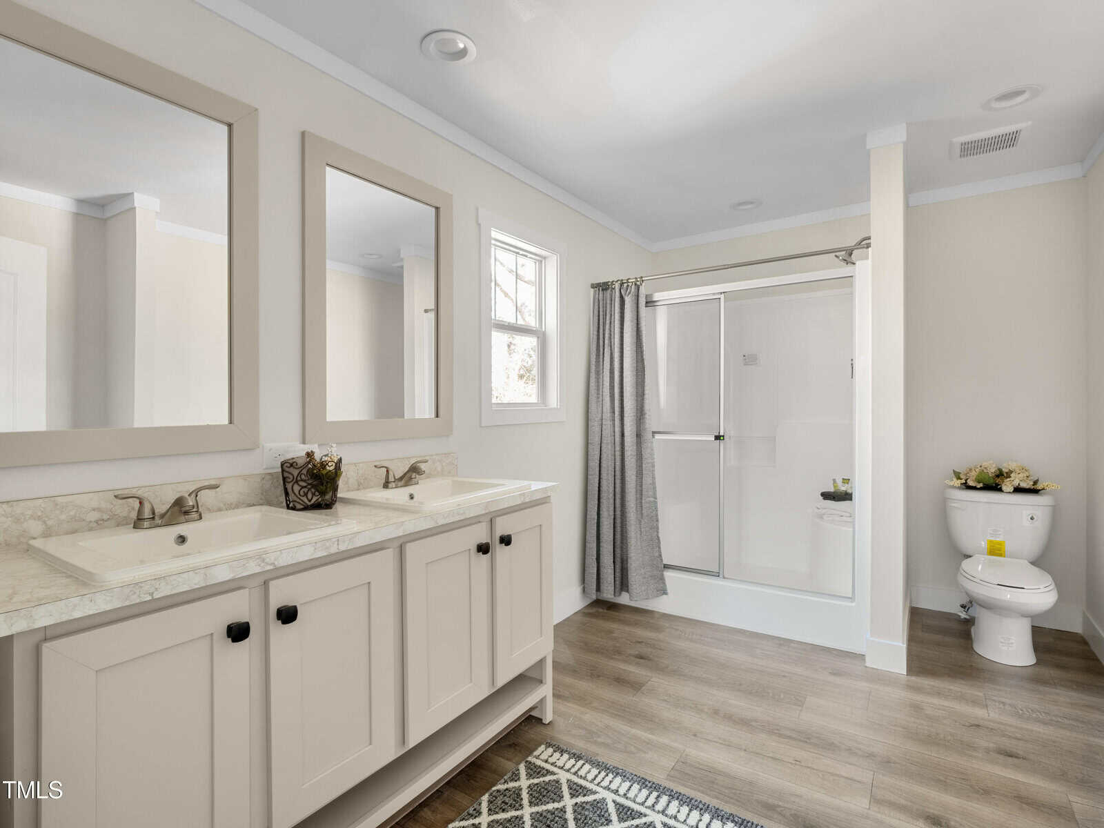 A modern bathroom features a white double sink vanity with matching mirrors, a glass-enclosed shower, and a white toilet. The space is illuminated by a small window above the sinks. The room has light walls and wood-look flooring, with a patterned rug near the vanity.
