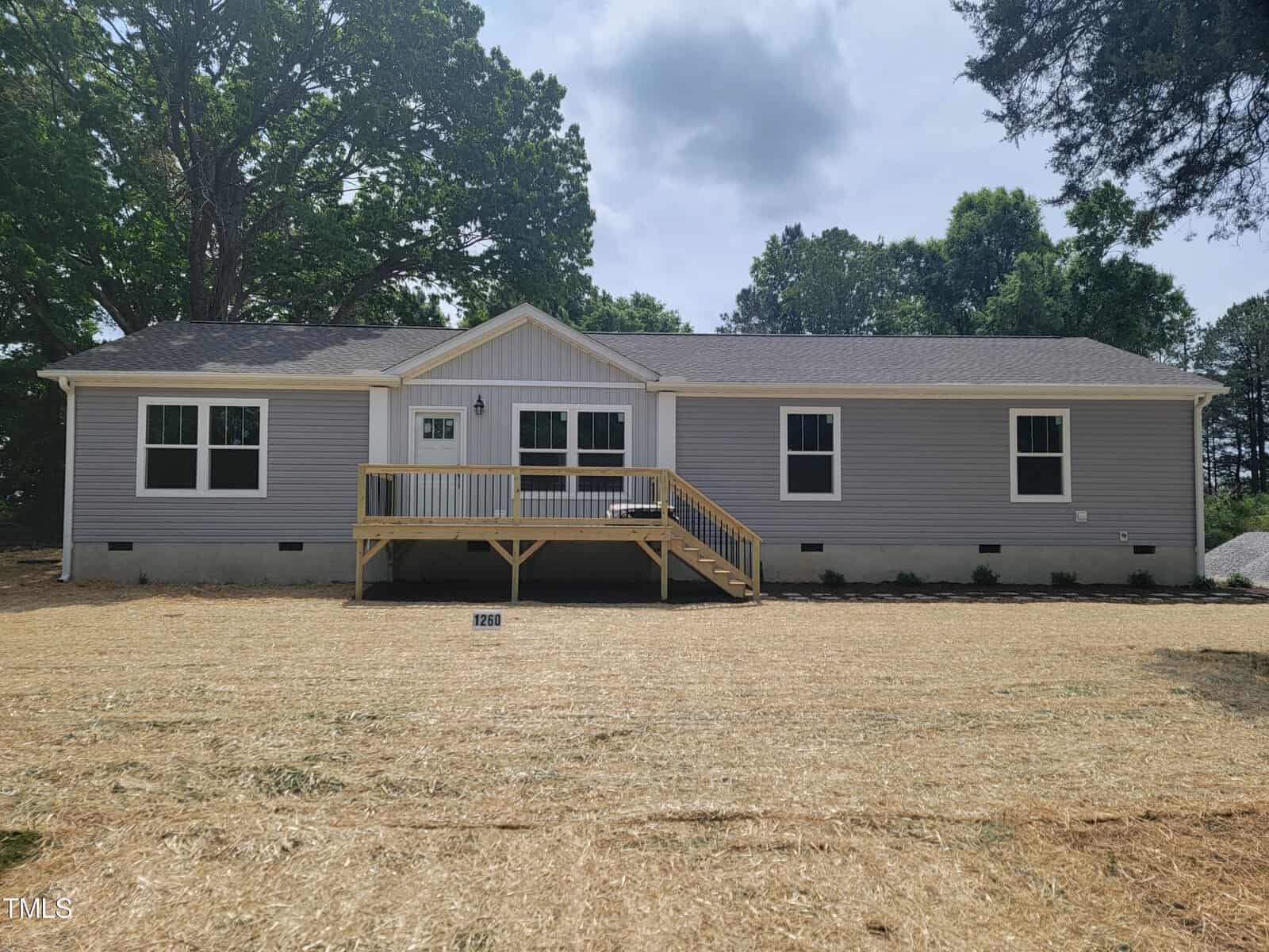 New single-story gray house located at 1260 Warrenton Road, Henderson NC, with a large front deck, surrounded by a freshly seeded lawn, under a clear sky. The house features double