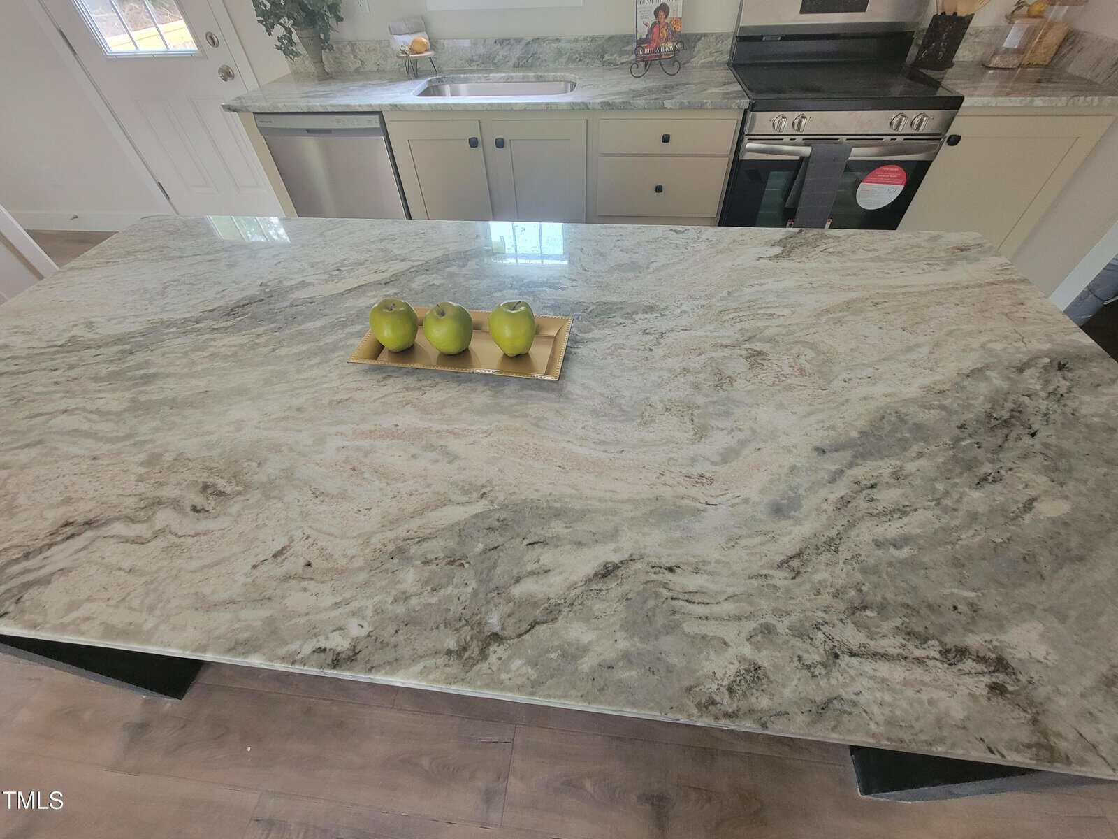 A modern kitchen with a large marble island featuring a green bowl holding three apples. the background shows white cabinets, a stove, and minimal decor.