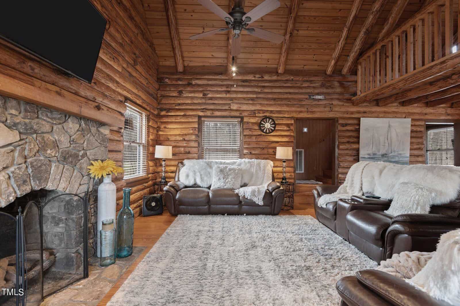 A cozy log cabin living room with a stone fireplace, wooden walls, a plush white rug, and leather furniture. decor includes a ceiling fan, wall clocks, and a vase of sunflowers.