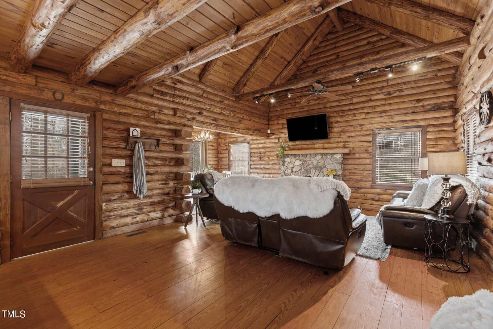 Cozy log cabin interior featuring a stone fireplace, wooden walls and floors, plush furniture, and a mounted flat-screen tv. decor includes rustic accents and elegant lighting.
