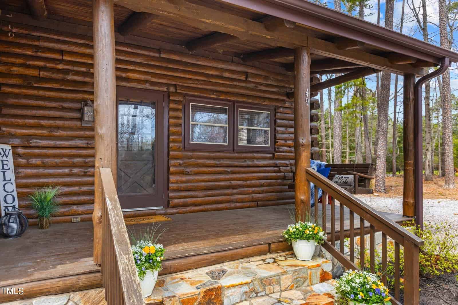A cozy log cabin with a welcoming sign above the door, surrounded by a wooded area. the front features a stone staircase, flowers in pots, and a porch with a bench.