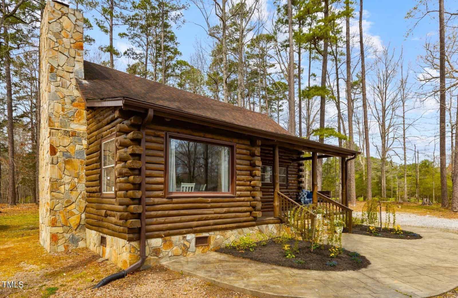 A rustic log cabin with a large stone chimney, nestled among tall pine trees, featuring a covered porch and a clear blue sky overhead.