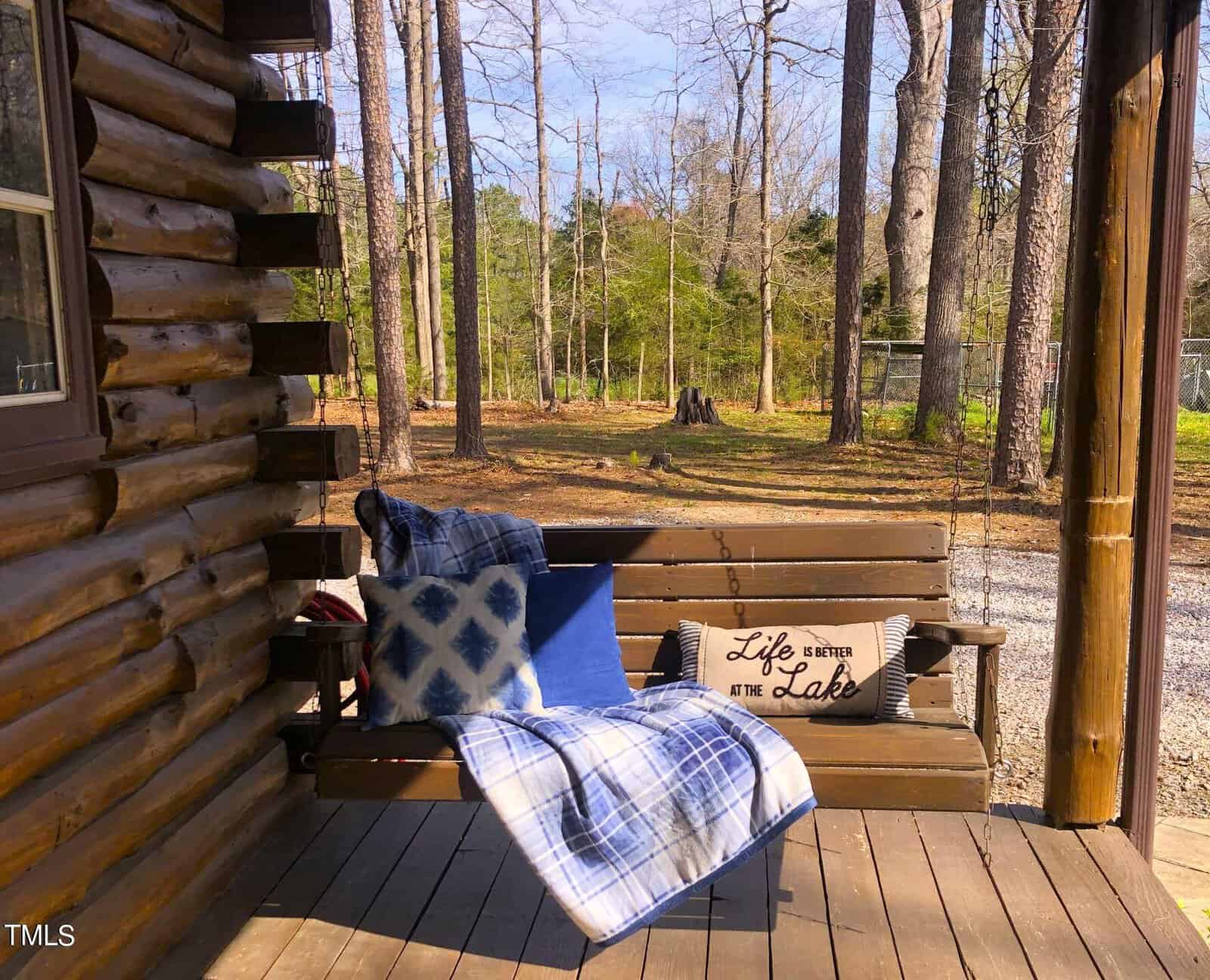 A cozy porch of a log cabin featuring a wooden bench with cushions and a "life is better at the lake" sign, overlooking a tranquil, wooded area.