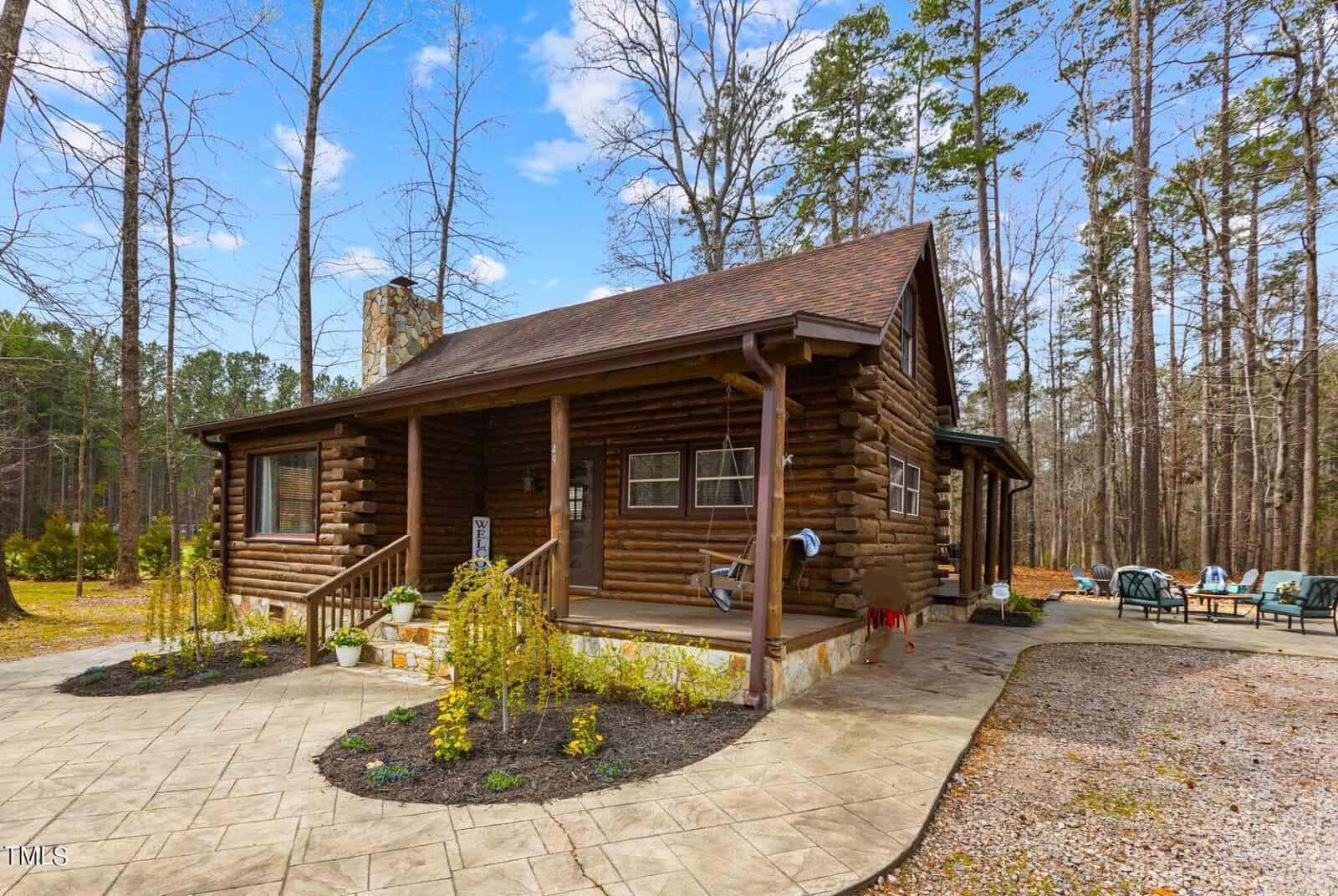 A log cabin surrounded by pine trees, located at 49 Pebble Hill Lane in Henderson NC, features a curved pathway and a small garden area. Outdoor furniture is set up on a patio to the