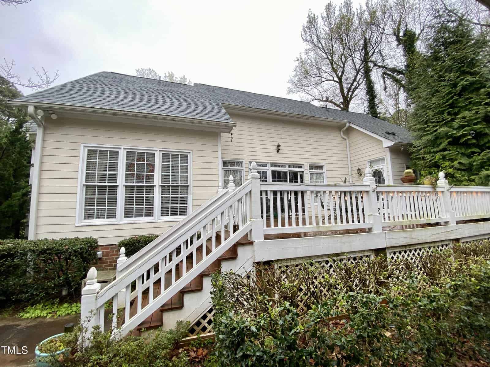 A beige single-story house with large white frame windows, a wooden deck with railing and stairs, and surrounded by greenery on a cloudy day.