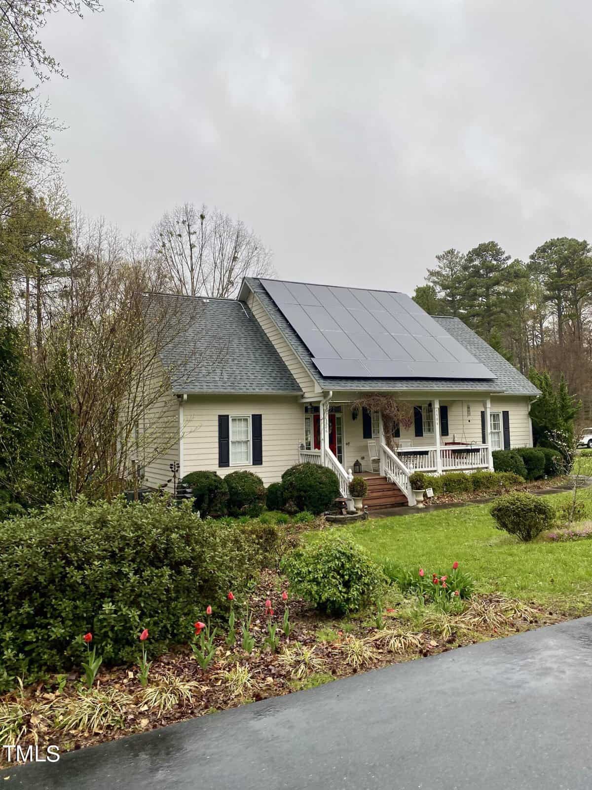 A single-story beige house with a dark roof featuring solar panels, a front porch with white railings, surrounded by greenery and red flowers, under a cloudy sky.