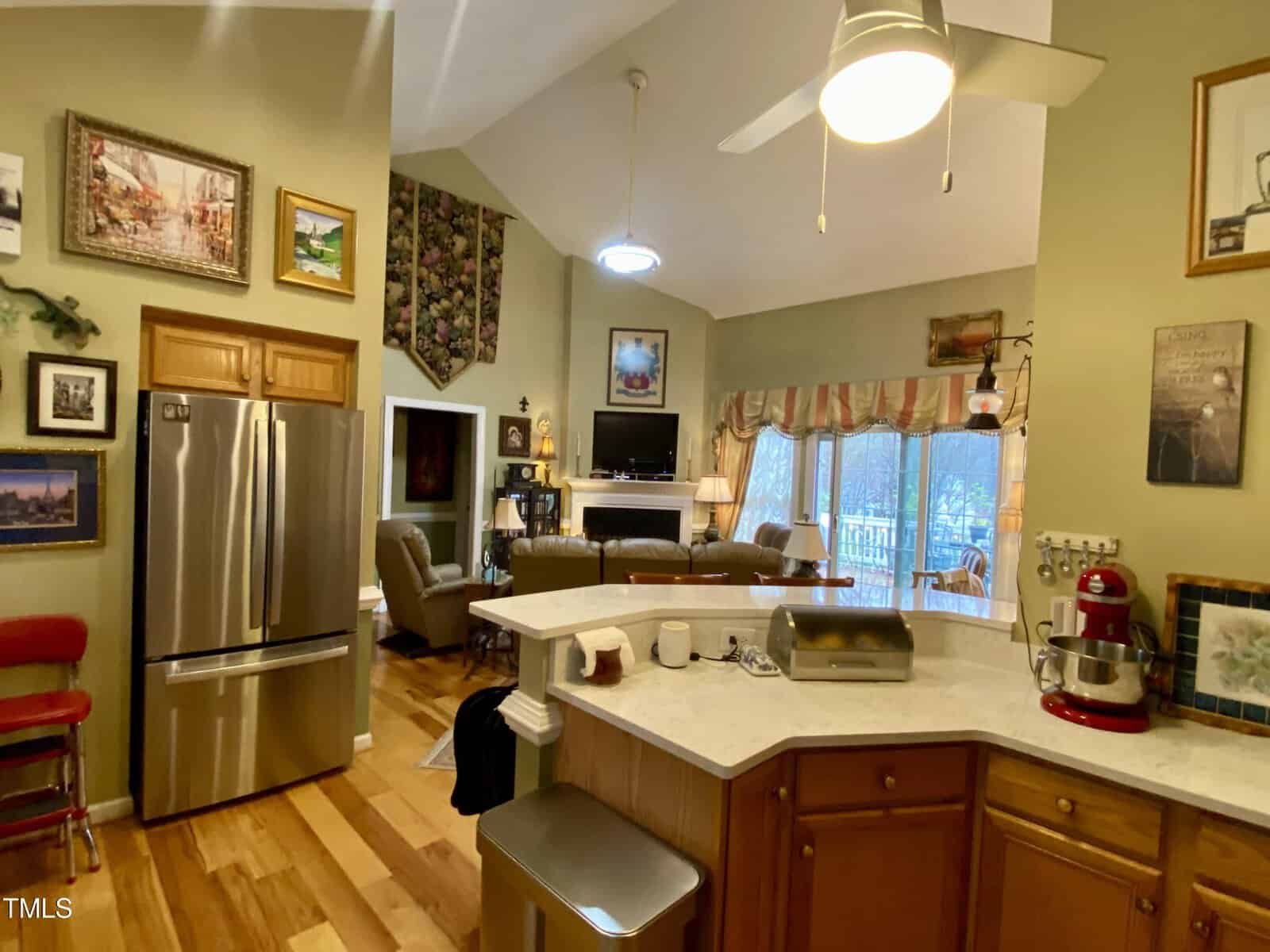 A cozy, well-lit kitchen opening into a living area, featuring stainless steel appliances, wooden cabinetry, and hardwood floors. the room includes a breakfast bar and a view to the outdoors through a large window with curtains.