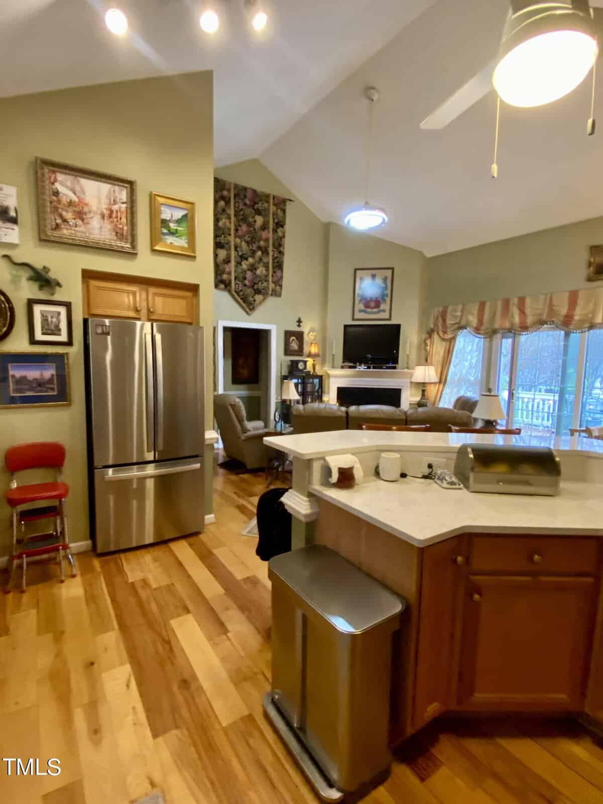 A cozy, well-lit kitchen with wooden floors, green walls, and modern appliances including a stainless steel refrigerator. the room extends into a living area with ample natural light.