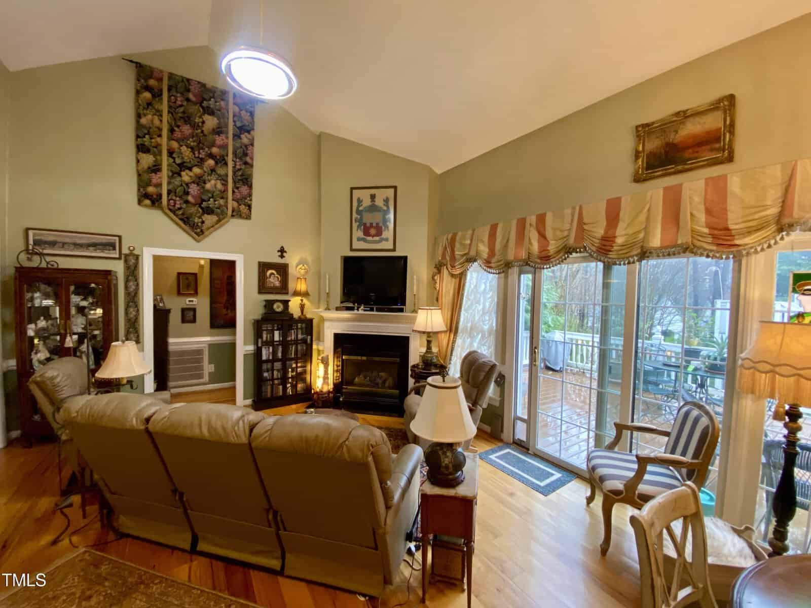 Cozy living room with high ceilings, featuring tan sofas, a fireplace, and traditional decor. wall arts and a dining area visible. large windows offer a view of greenery outside.