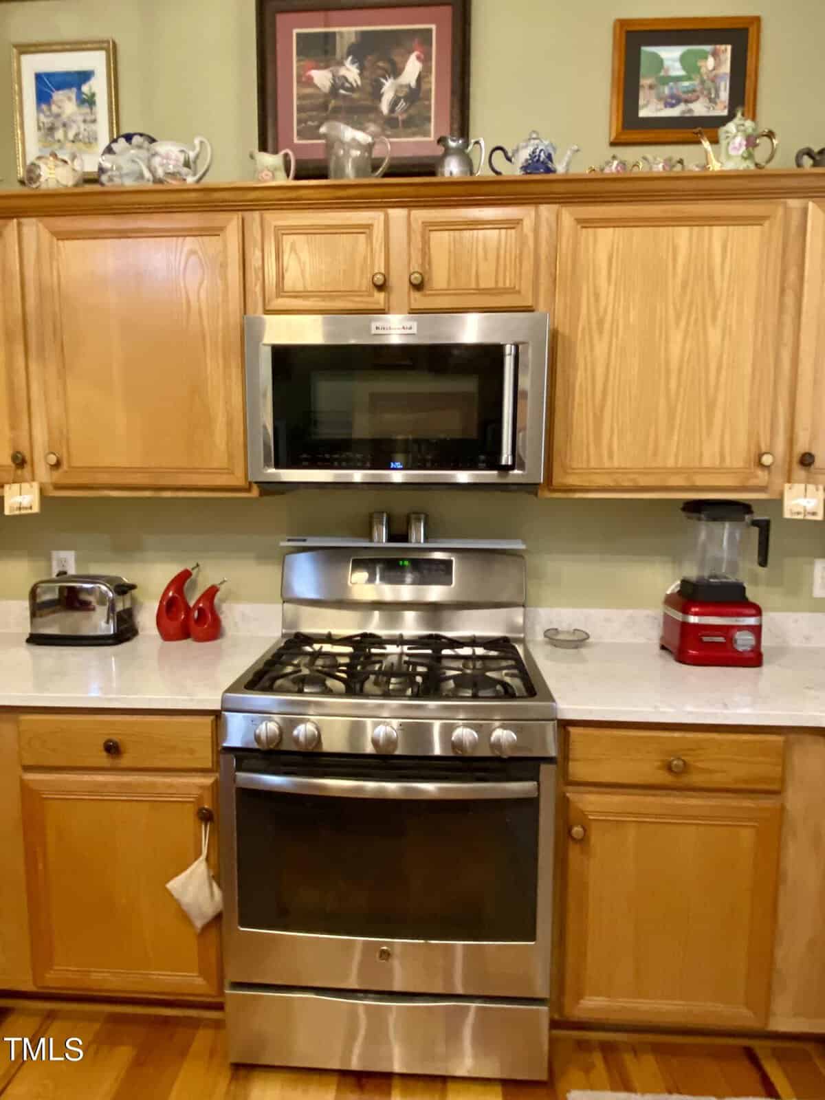 A cozy kitchen featuring wooden cabinets, stainless steel oven with gas stove, microwave above, and small red appliances on a white countertop. decorative items rest on top of the cabinets.