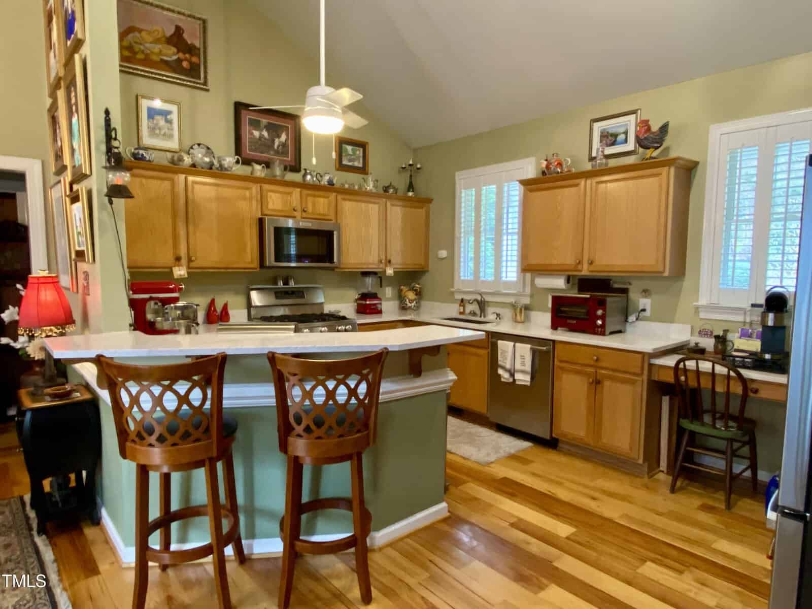 A cozy kitchen featuring wooden cabinetry, a central island with high stools, hardwood floors, various kitchen appliances, and decorative items. light from windows brightens the space.