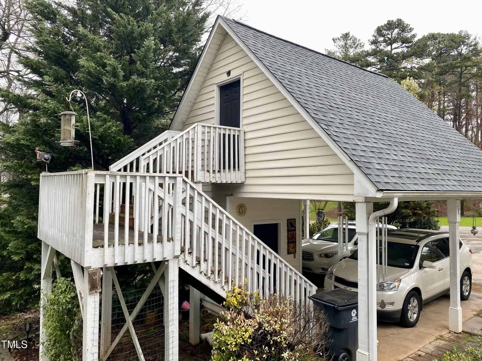 A white two-story house with a slanted roof and an elevated wooden deck with stairs leading up to it. there are parked cars under the deck, surrounded by trees and shrubbery.