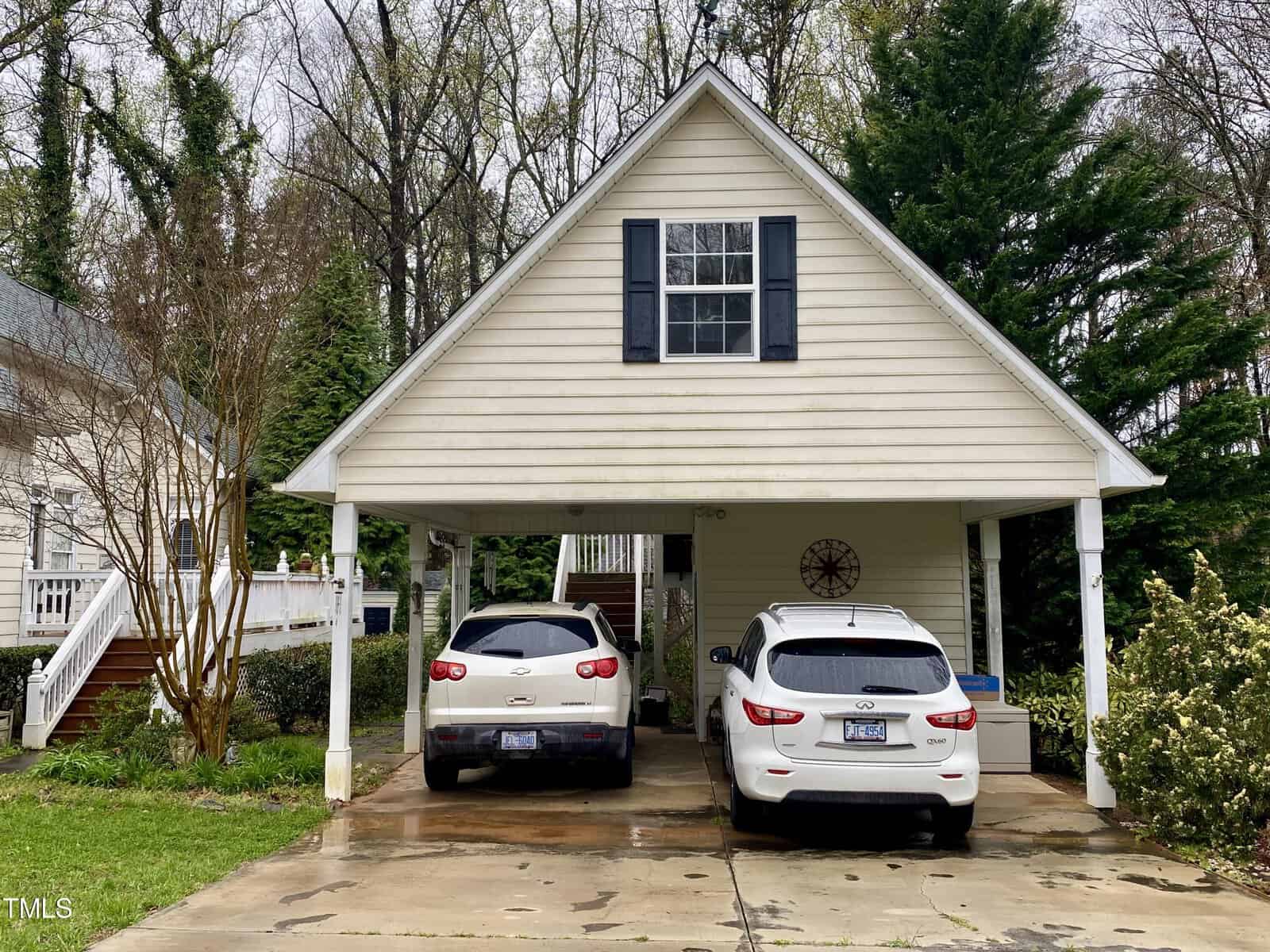 A two-story home with a carport sheltering two cars, surrounded by lush greenery and a wet driveway, indicating recent rainfall.