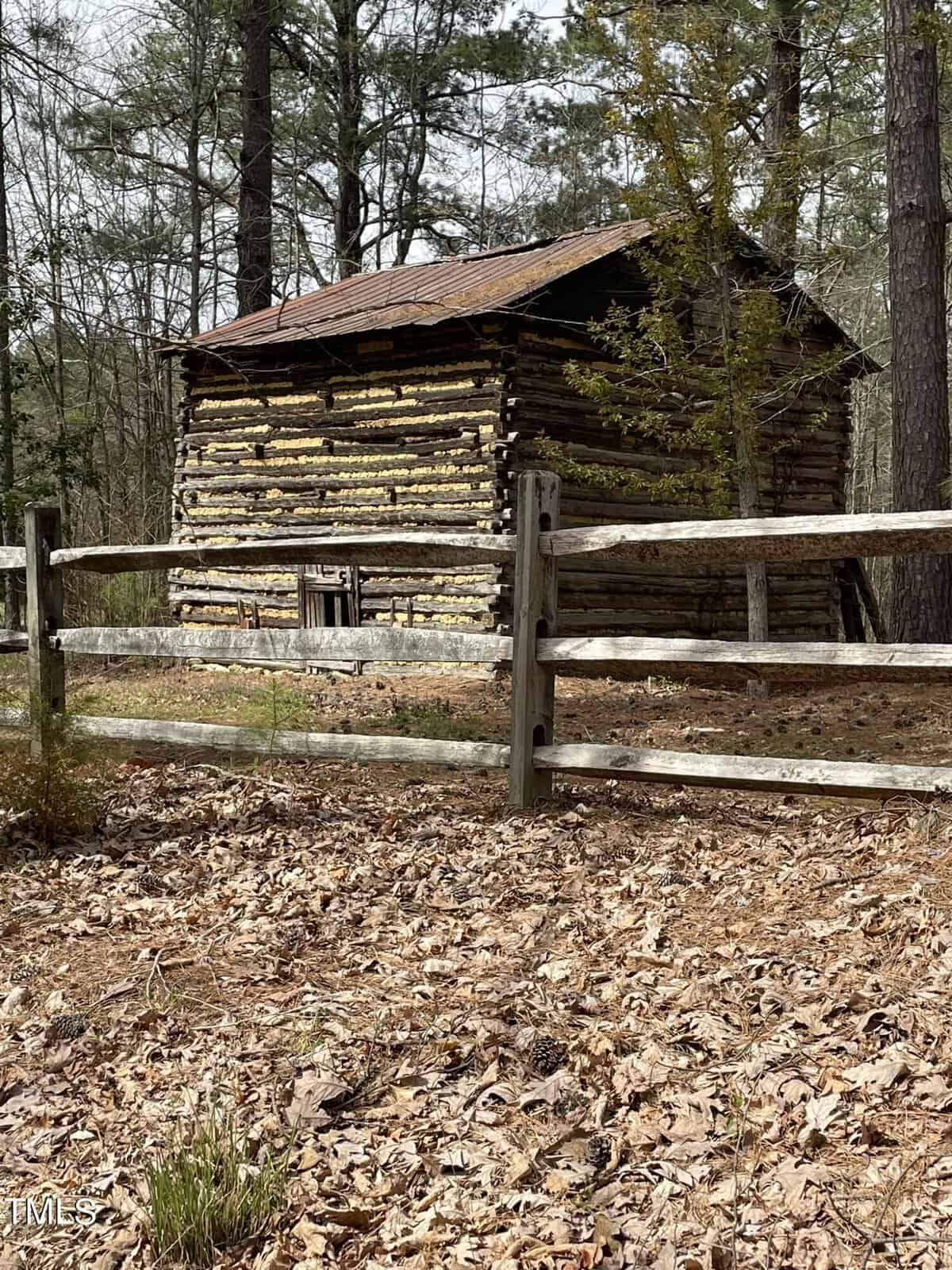 A rustic log cabin with a weathered wooden roof is situated in a forest. It is surrounded by tall trees and a wooden fence in the foreground. The ground is covered with fallen leaves, indicating a season of autumn or early spring.