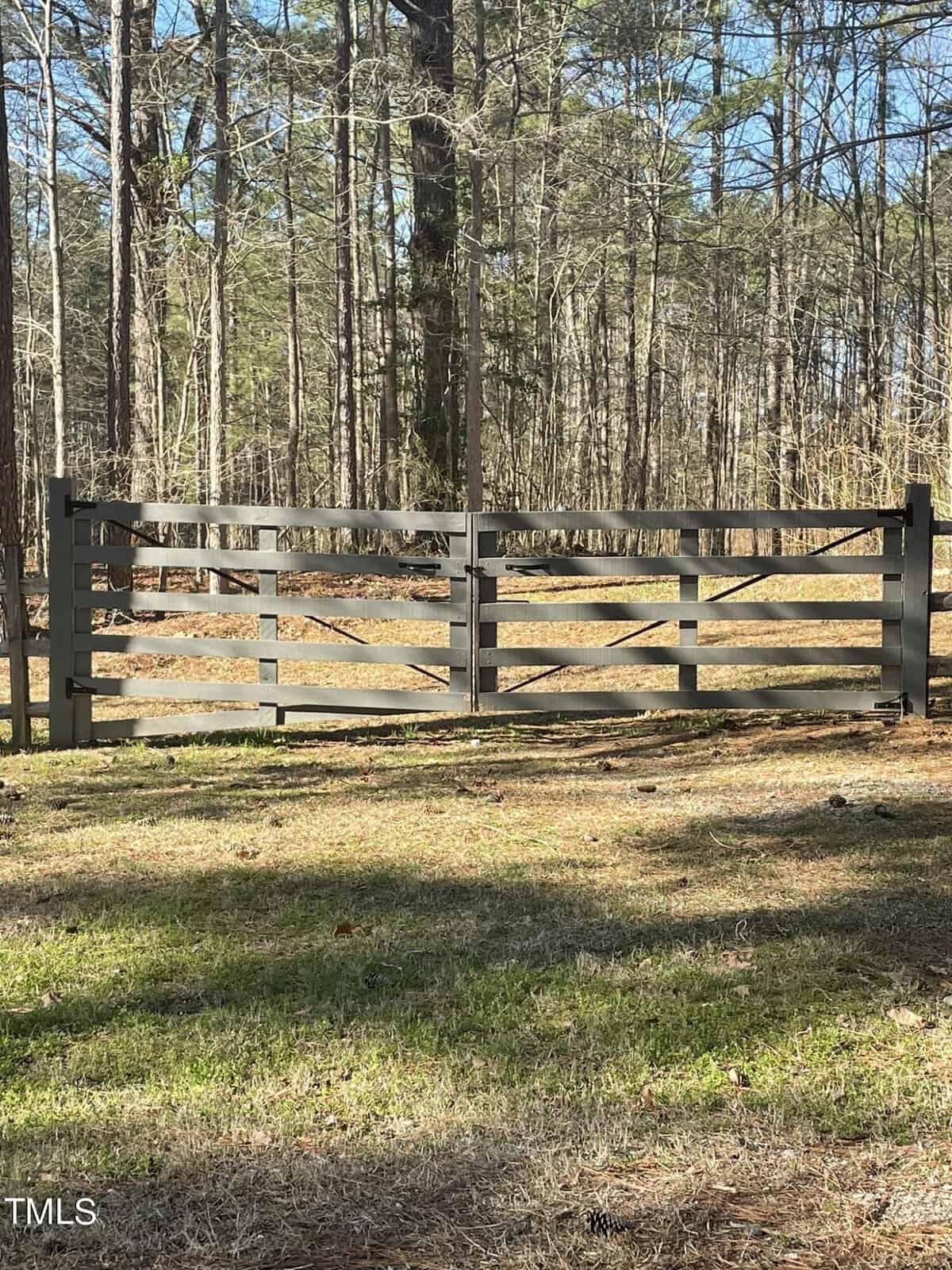 A wooden double gate stands in a forest setting near 1412 Buchanan Road, with tall trees in the background. Sunlight casts shadows on the grass-covered ground, creating a serene and natural scene reminiscent of a countryside manson.