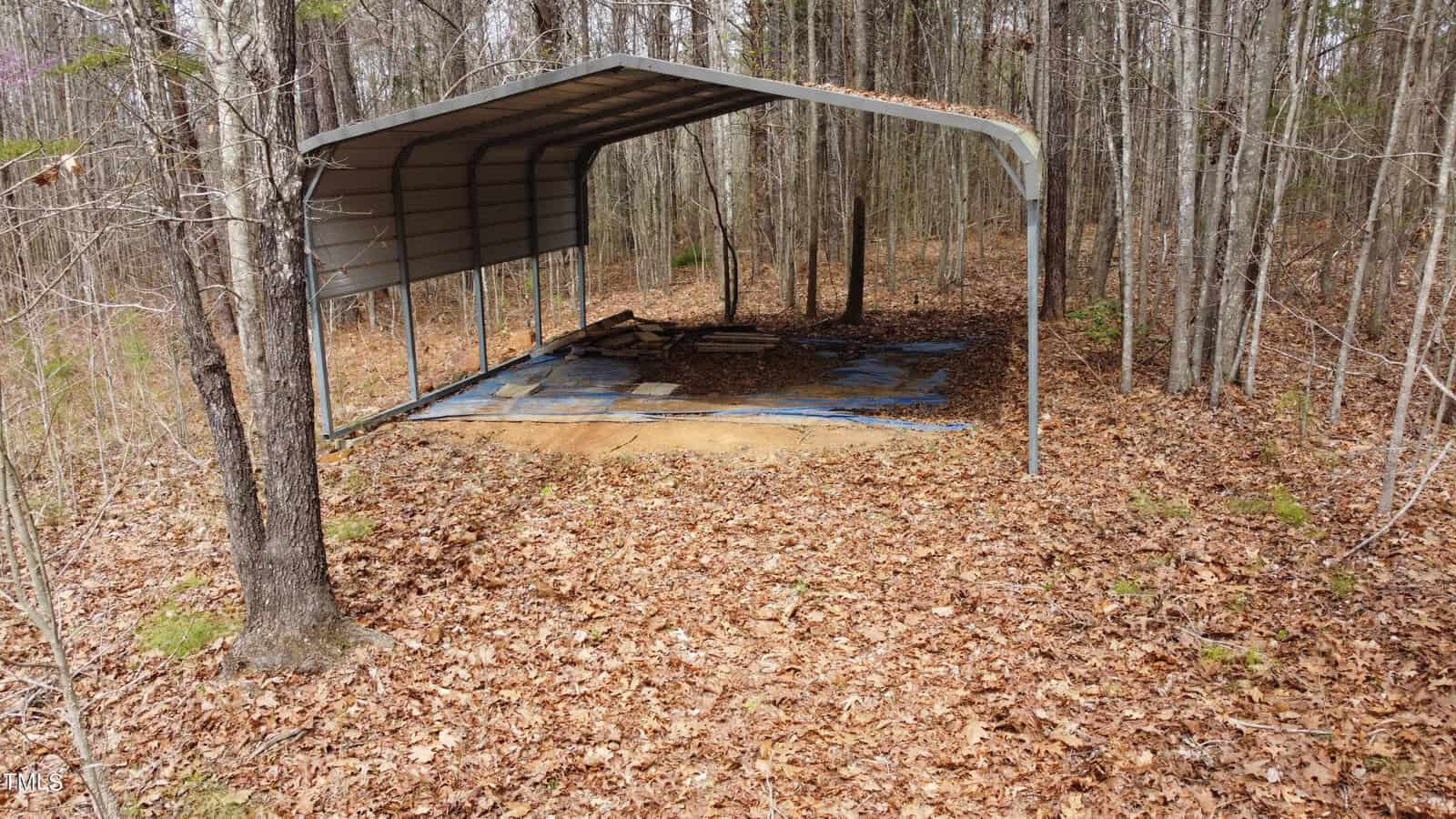 A metal carport stands in a forested area, surrounded by trees and dry leaves on the ground. The carport covers some wooden planks and a concrete base, with sunlight filtering through the branches.