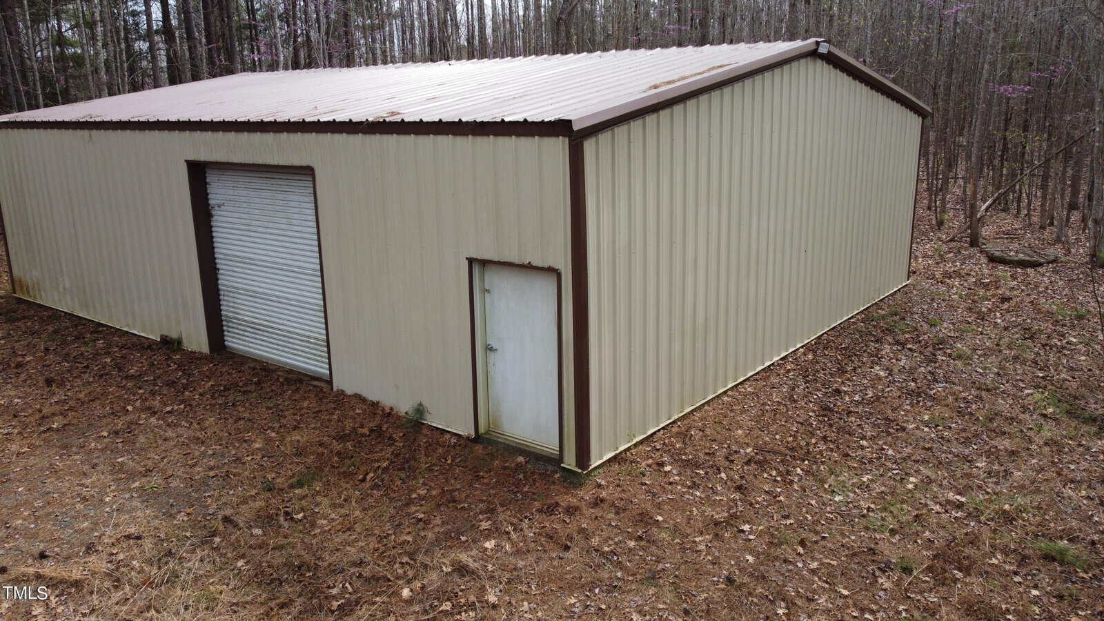 A large, tan metal building with a brown roof, featuring a roll-up door and a smaller entry door, is situated in a wooded area. Fallen leaves cover the ground around the structure. There are tall trees surrounding the building.