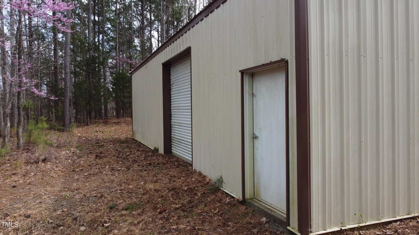 A beige metal building with a large garage door and a smaller side door stands on a wooded plot, surrounded by pine trees and pink flowering trees. Brown leaves cover the ground around the structure.