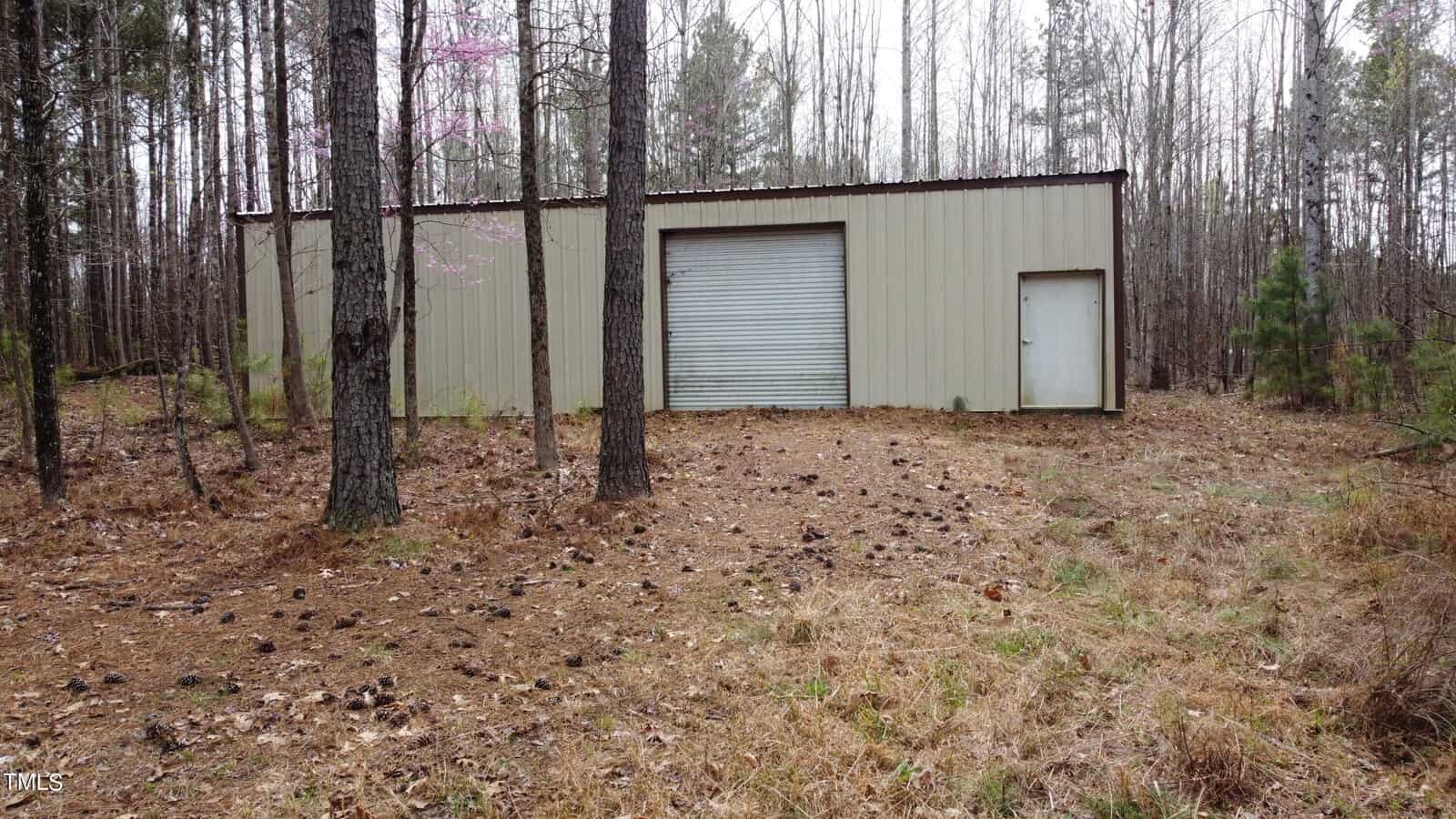 A beige metal storage building with a large roll-up door is surrounded by tall trees and a forest floor covered with pine needles. Sparse trees and a partially blooming pink tree are visible in the background.