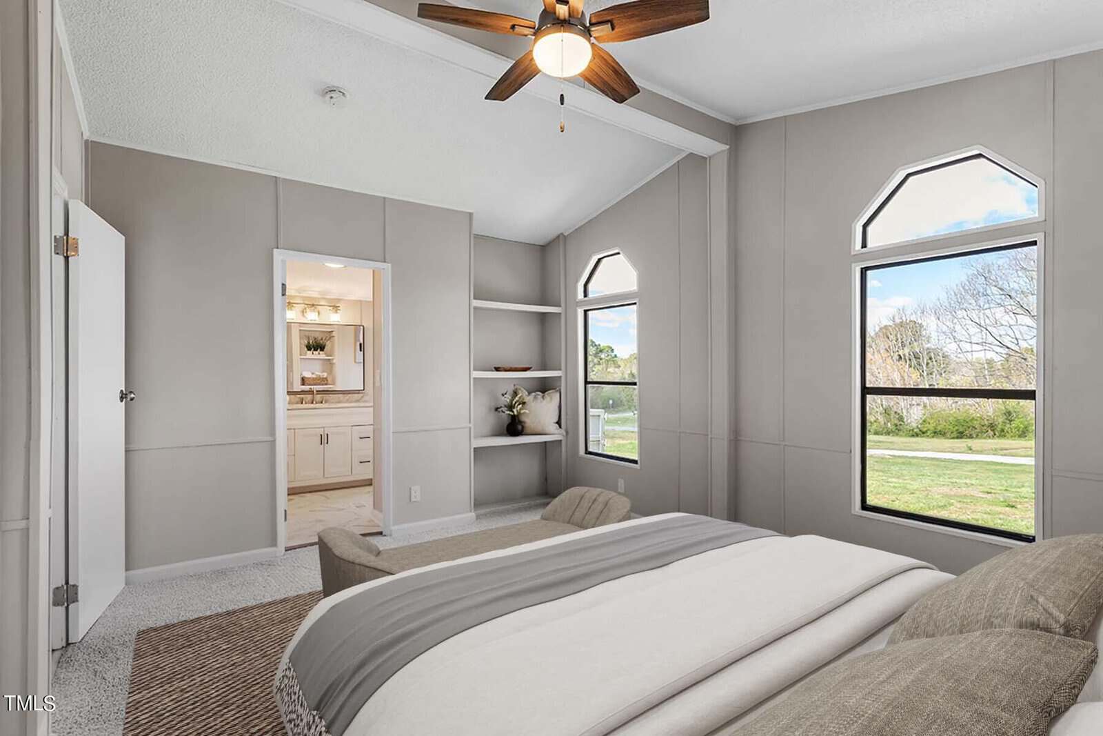 Modern bedroom with a large bed, gray walls, vaulted ceiling with a ceiling fan, and two large windows showing a sunny rural view. there are built-in shelves and a doorway leading to another room.