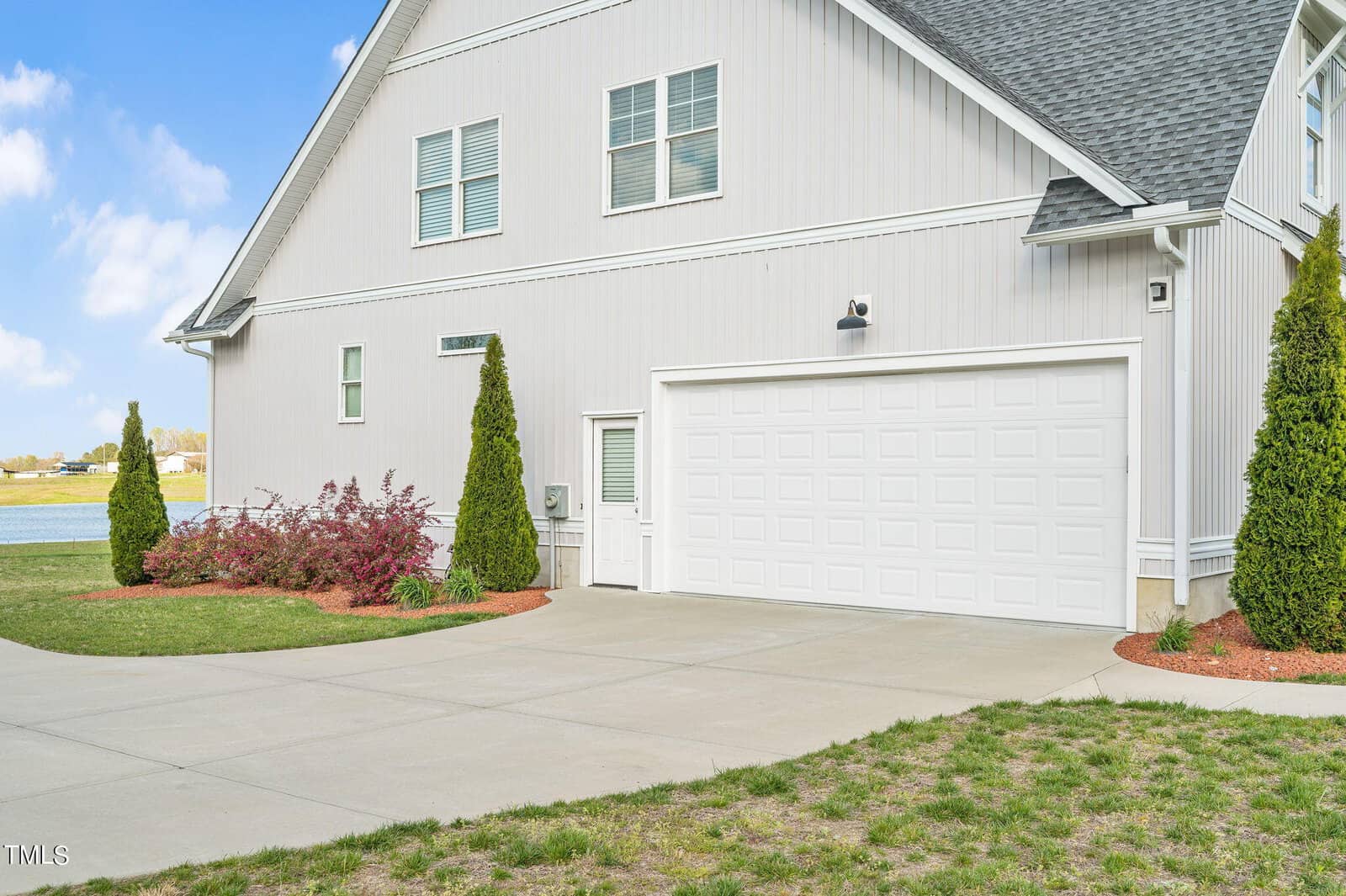 A modern white house with a large garage and paved driveway. The garden features neatly trimmed bushes and small trees. A clear blue sky and a body of water can be seen in the background.