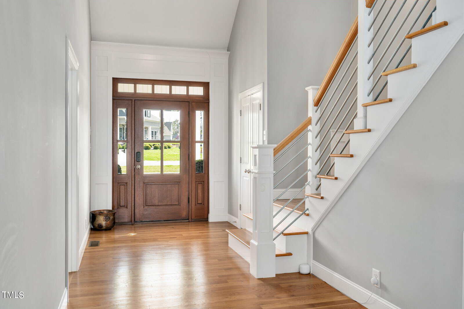 A bright entryway featuring a wooden front door with glass panels. The space has light-colored walls and hardwood floors. A staircase with white railings and wooden handrails is on the right. Sunlight streams in, illuminating the room.