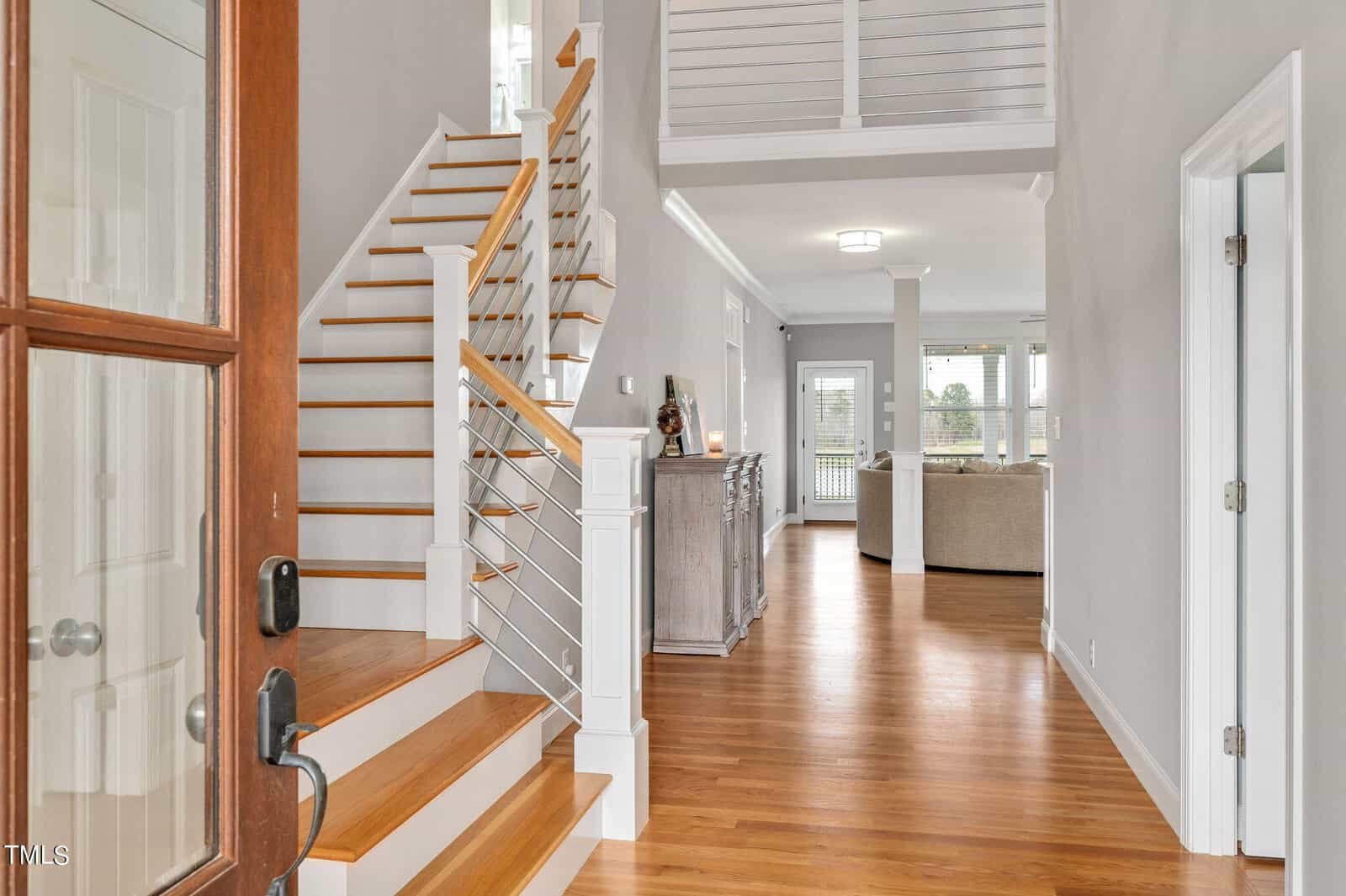 A modern home interior featuring a polished wooden staircase with sleek metal railings. The hallway has hardwood floors, gray walls, and opens to a bright living area with large windows and a view of a sofa and a sideboard.