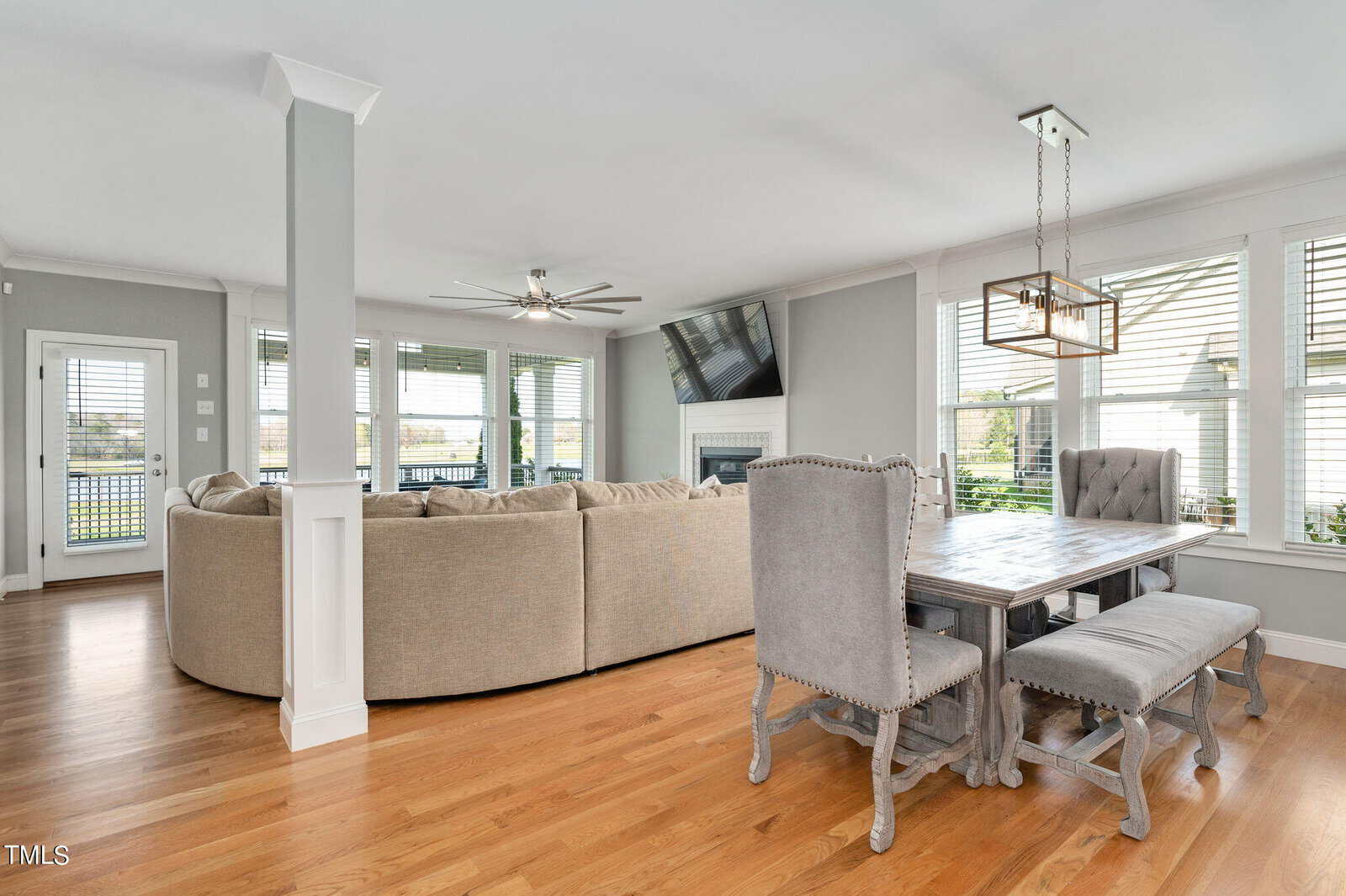 Bright living and dining area with a large beige sectional sofa facing a flat-screen TV above a fireplace. Grey dining chairs surround a rustic wooden table. Windows with white shutters let in natural light, highlighting the hardwood floors.