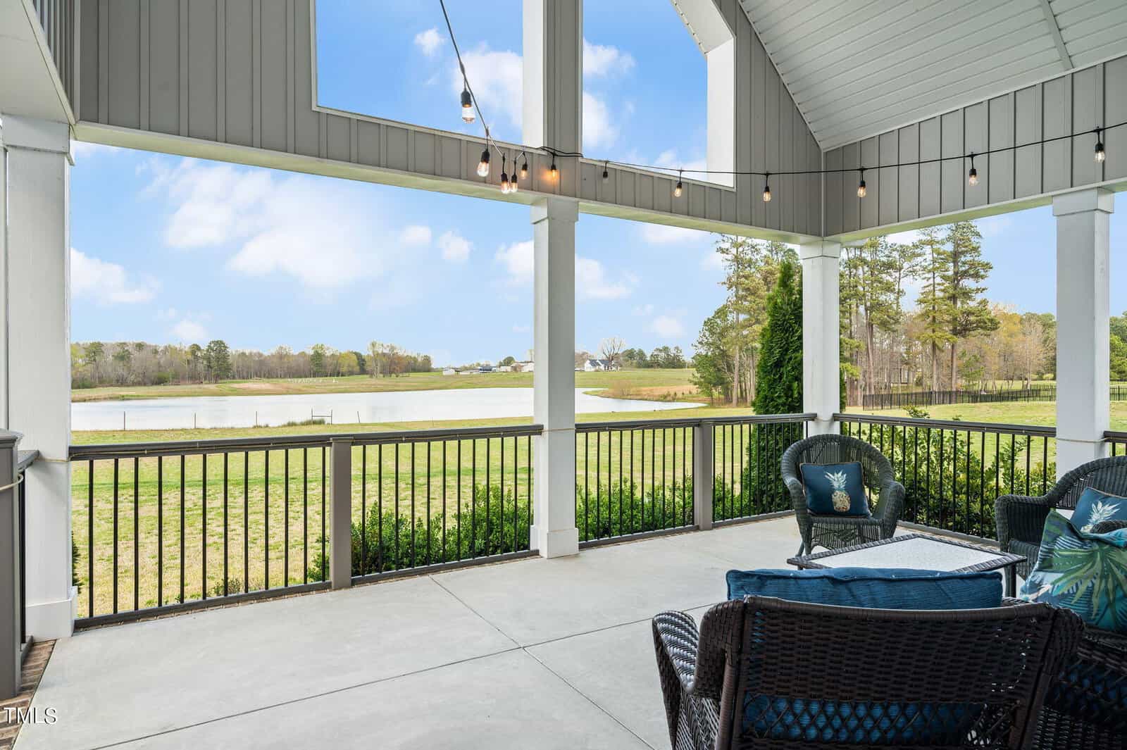 A spacious patio with seating overlooks a scenic body of water. The area is decorated with string lights under a high ceiling, surrounded by a black railing, and offers views of a grassy landscape and trees in the distance under a blue, cloudy sky.