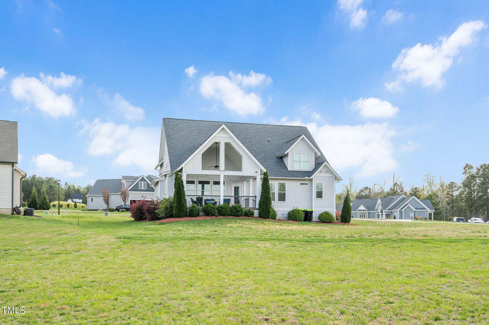 A charming white house with a gray roof and large porch, surrounded by a spacious green lawn. The sky is clear with scattered clouds. Multiple homes are visible in the background, set in a tranquil neighborhood.
