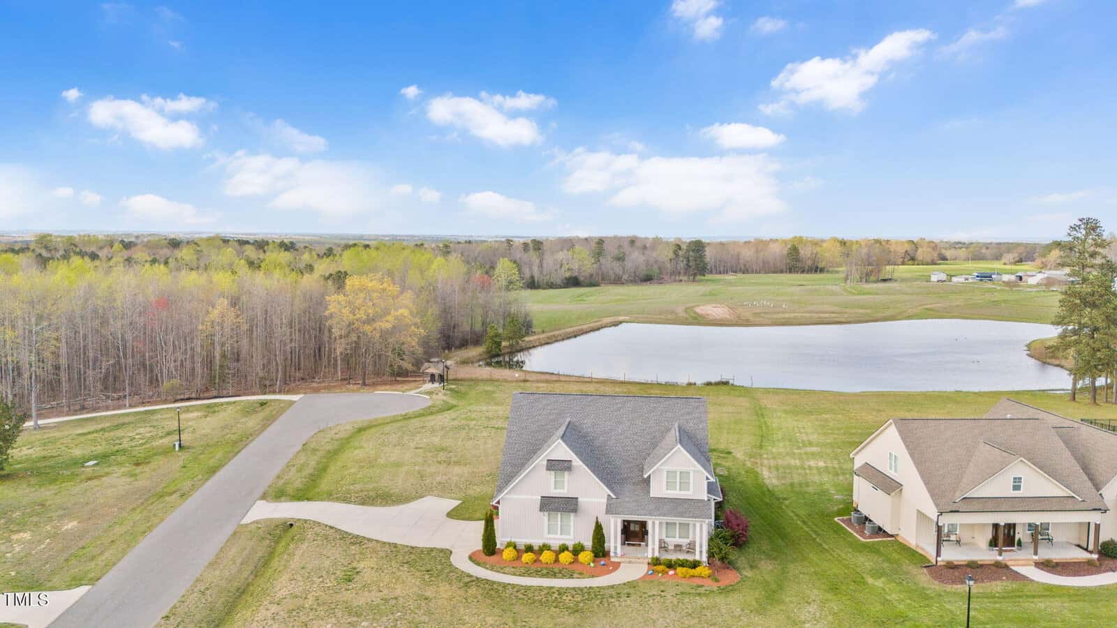 Aerial view of a suburban landscape featuring a white house with a neatly manicured lawn and garden. A pond and wooded area lie in the background under a blue sky with scattered clouds. Two other houses are visible nearby.