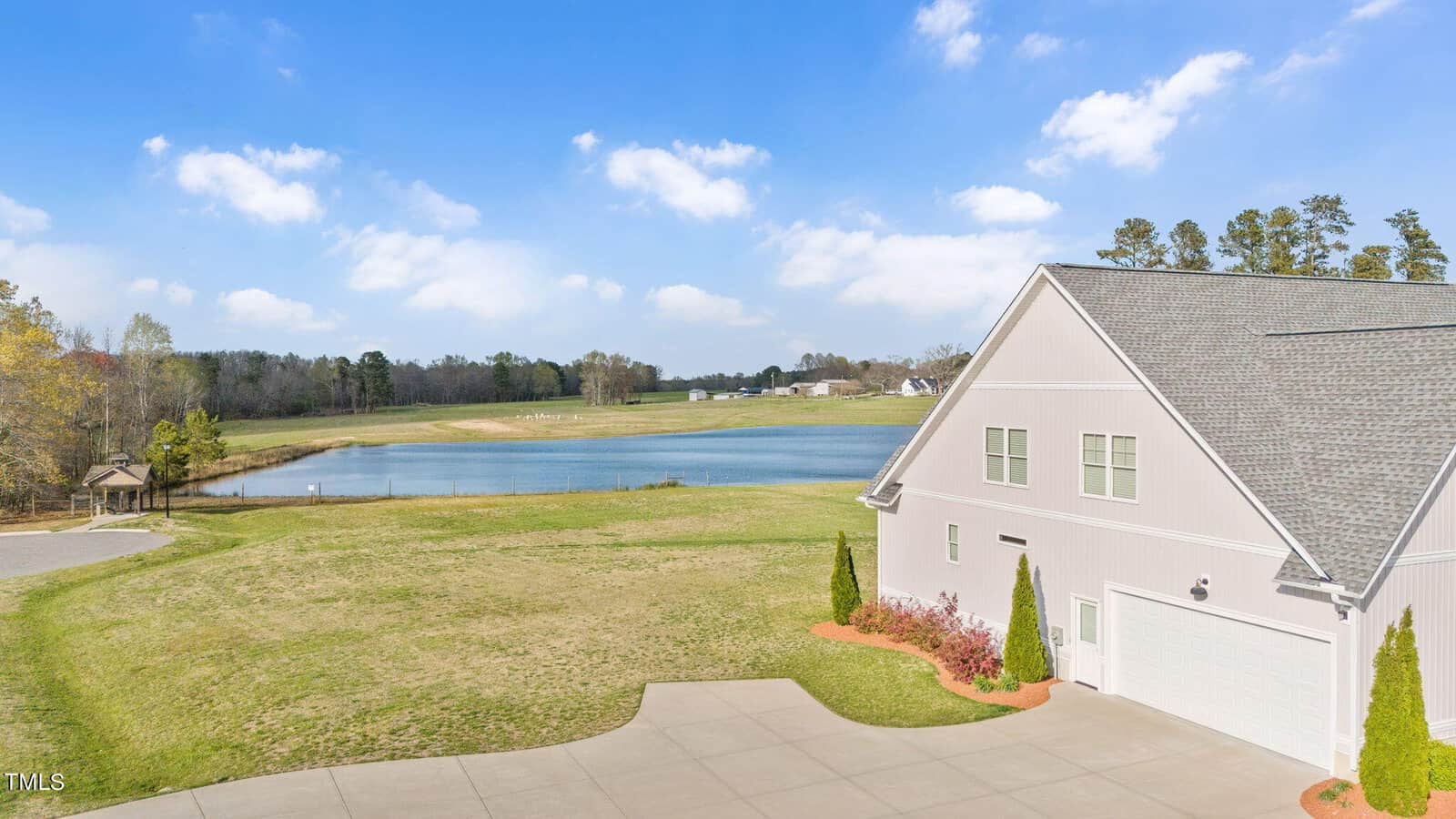 A serene landscape featuring a pond surrounded by grassy fields and scattered trees under a blue sky with clouds. In the foreground, a modern barn-shaped building with a paved driveway is visible.