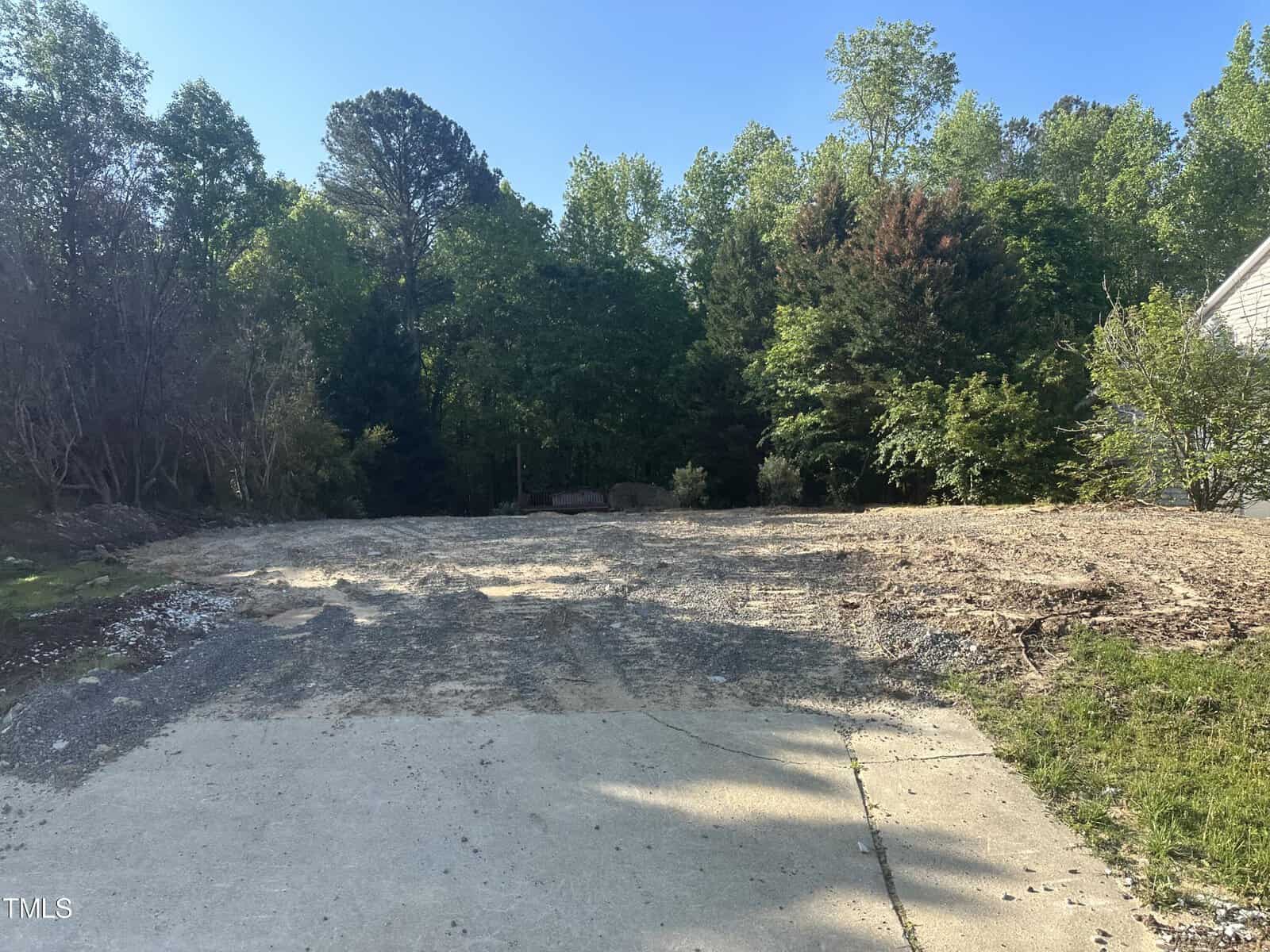 A cleared, empty lot with a concrete driveway surrounded by trees and greenery on a sunny day. The ground appears uneven with traces of construction or landscaping work.
