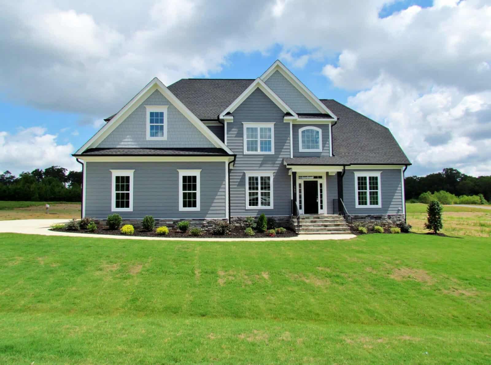 A large two-story gray house with white trim, multiple gables, and a dark roof sits on a spacious, well-manicured green lawn under a partly cloudy sky. Shrubs and small plants line the front of the house.