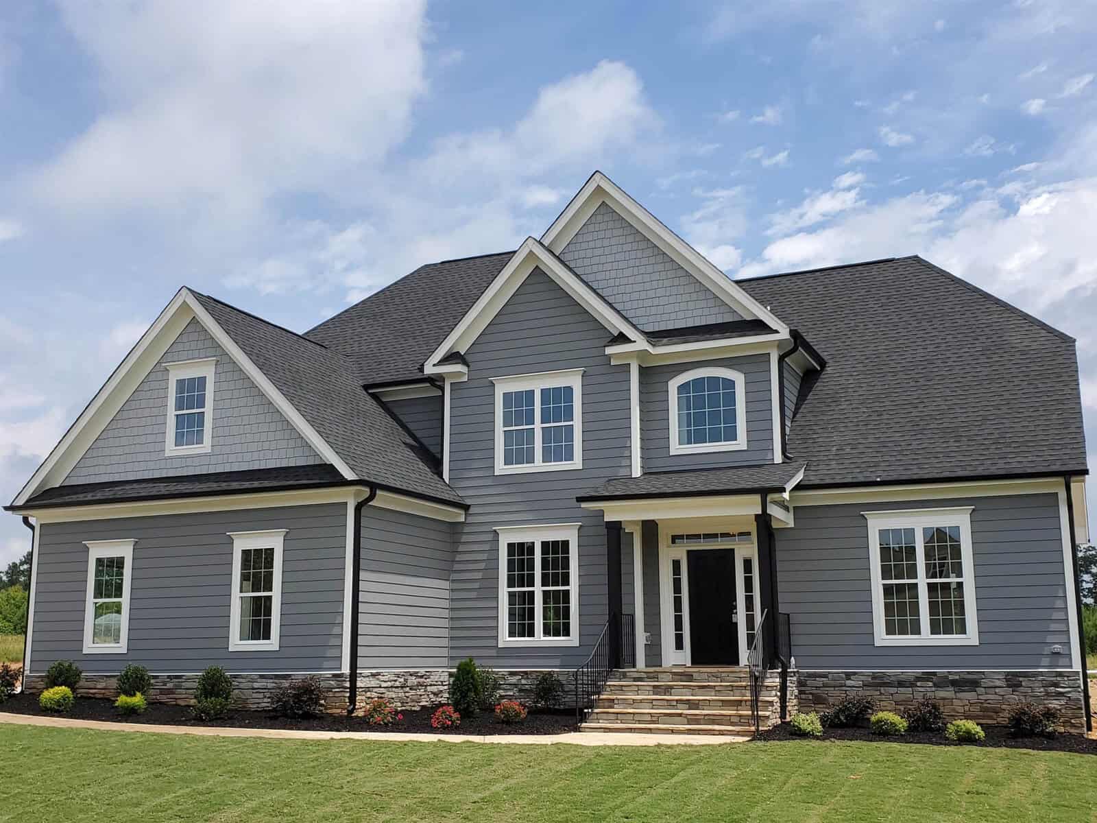 A modern two-story house with gray siding, white trim, and dark roof. The front features multiple gables, large windows, a black front door with glass panels, stone accents, and landscaped garden beds.