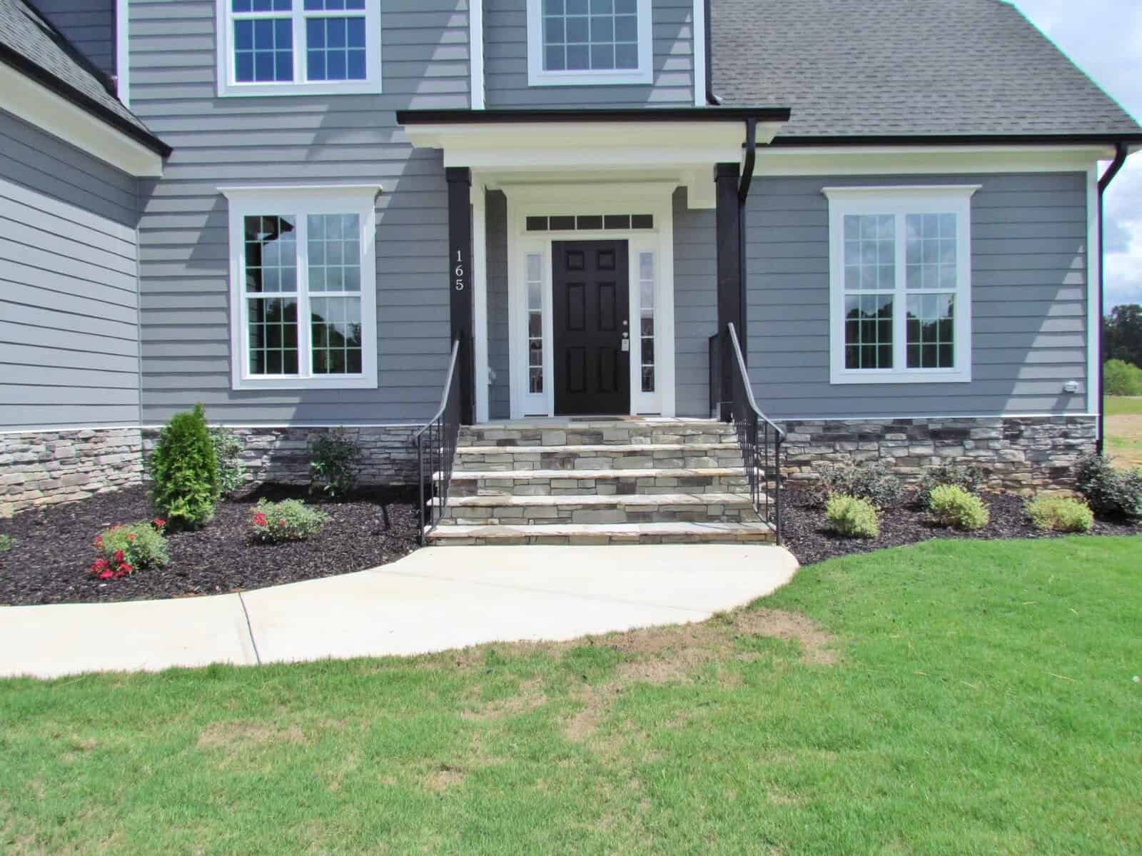 A modern gray house with white trim, stone accents, and a black front door. Stone steps lead to the entrance, bordered by metal railings and landscaped flower beds next to a green lawn.