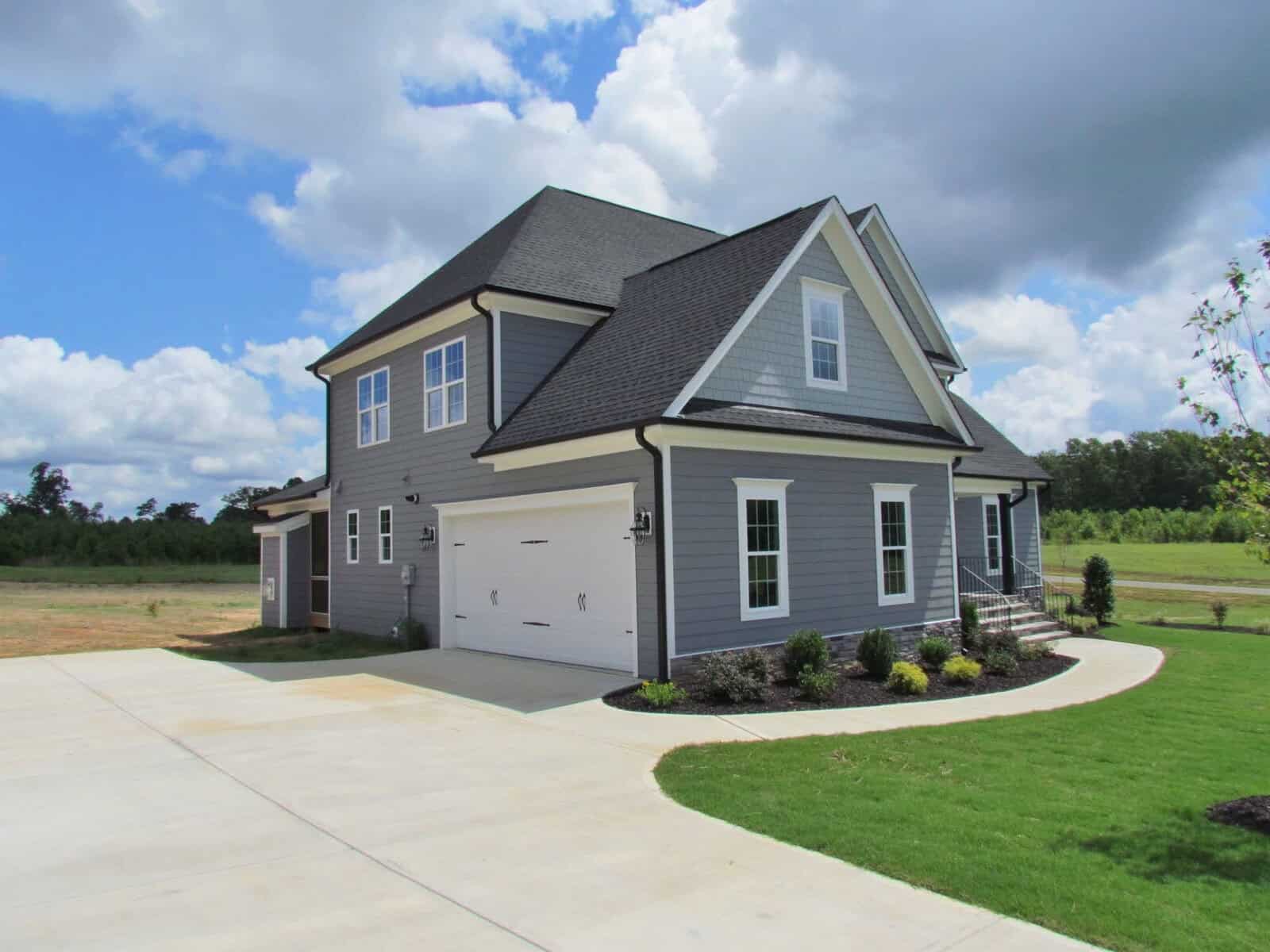 A modern two-story gray house with white trim, a double garage, and a manicured lawn sits under a partly cloudy sky with a concrete driveway leading up to it.