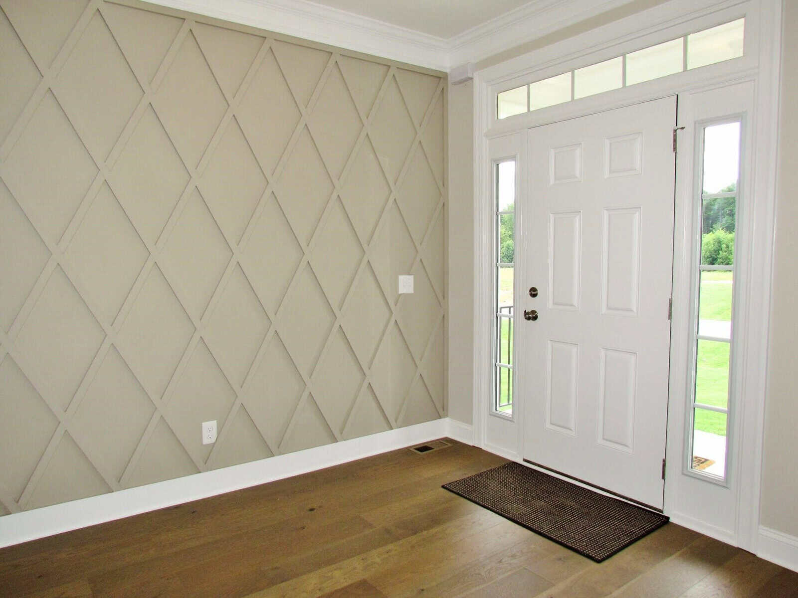 A bright entryway with a white front door, sidelights, transom window, wood floor, and a beige accent wall featuring a diamond pattern of raised trim. A small brown doormat sits in front of the door.
