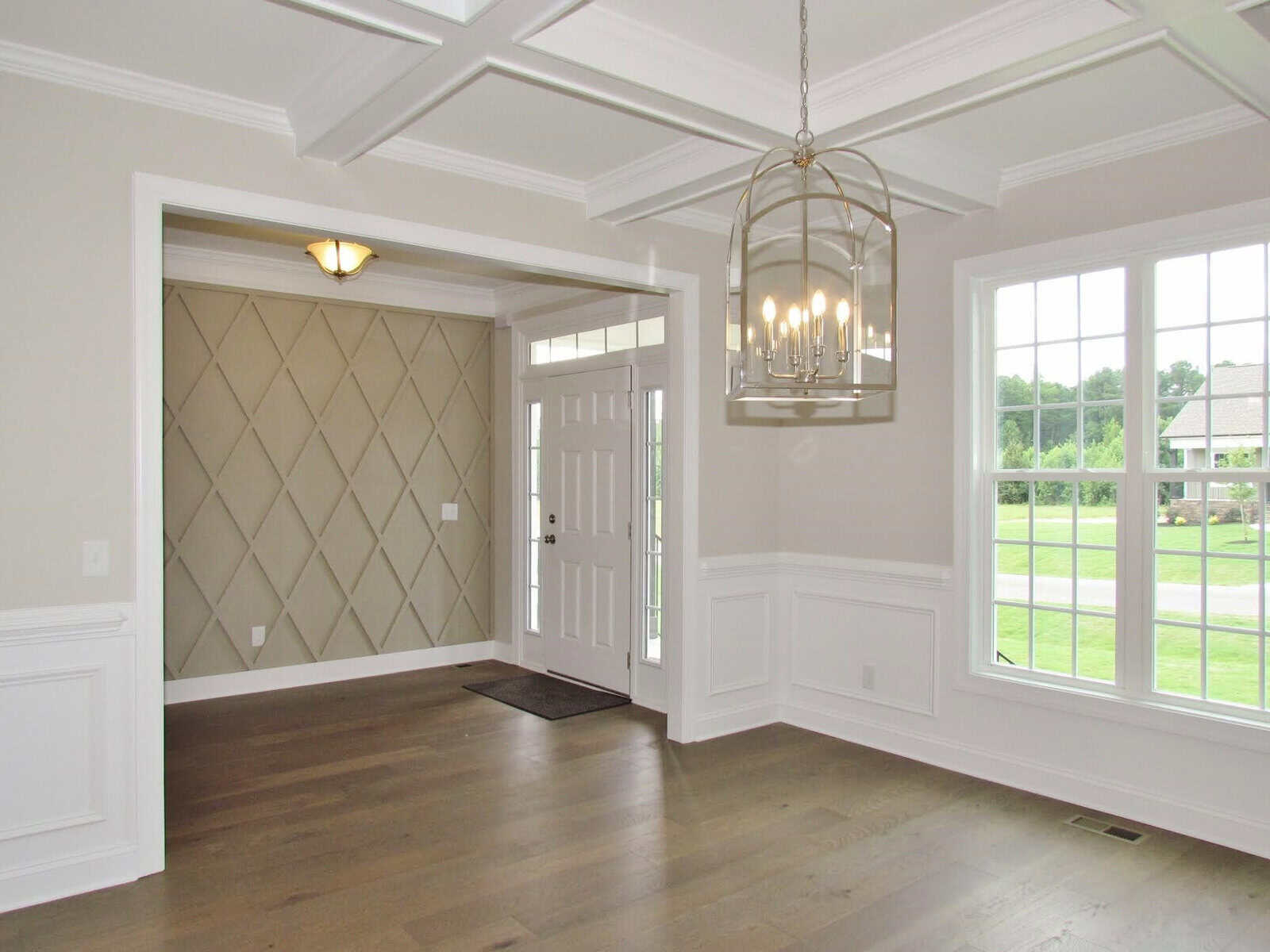 A bright, empty room with light wood flooring, white paneled walls, large windows, a coffered ceiling, a metallic chandelier, and a white front door with a diamond-pattern accent wall in the entryway.