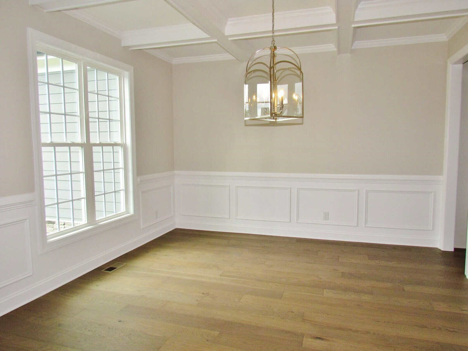 A bright, empty dining room with large windows, light beige walls, white wainscoting, a wooden floor, a coffered ceiling, and a gold chandelier hanging in the center.