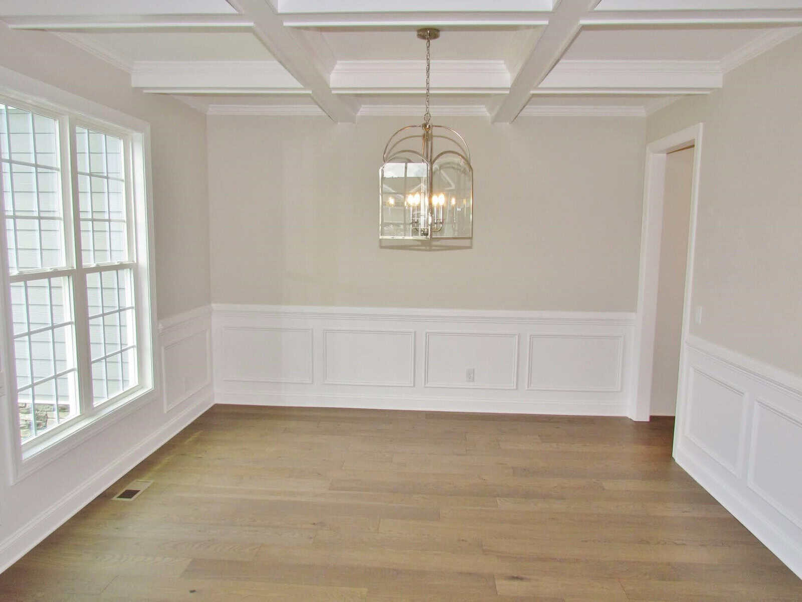 A bright, empty dining room with white wainscoting, beige walls, a coffered ceiling, a large window, wood floors, and a hanging chandelier.